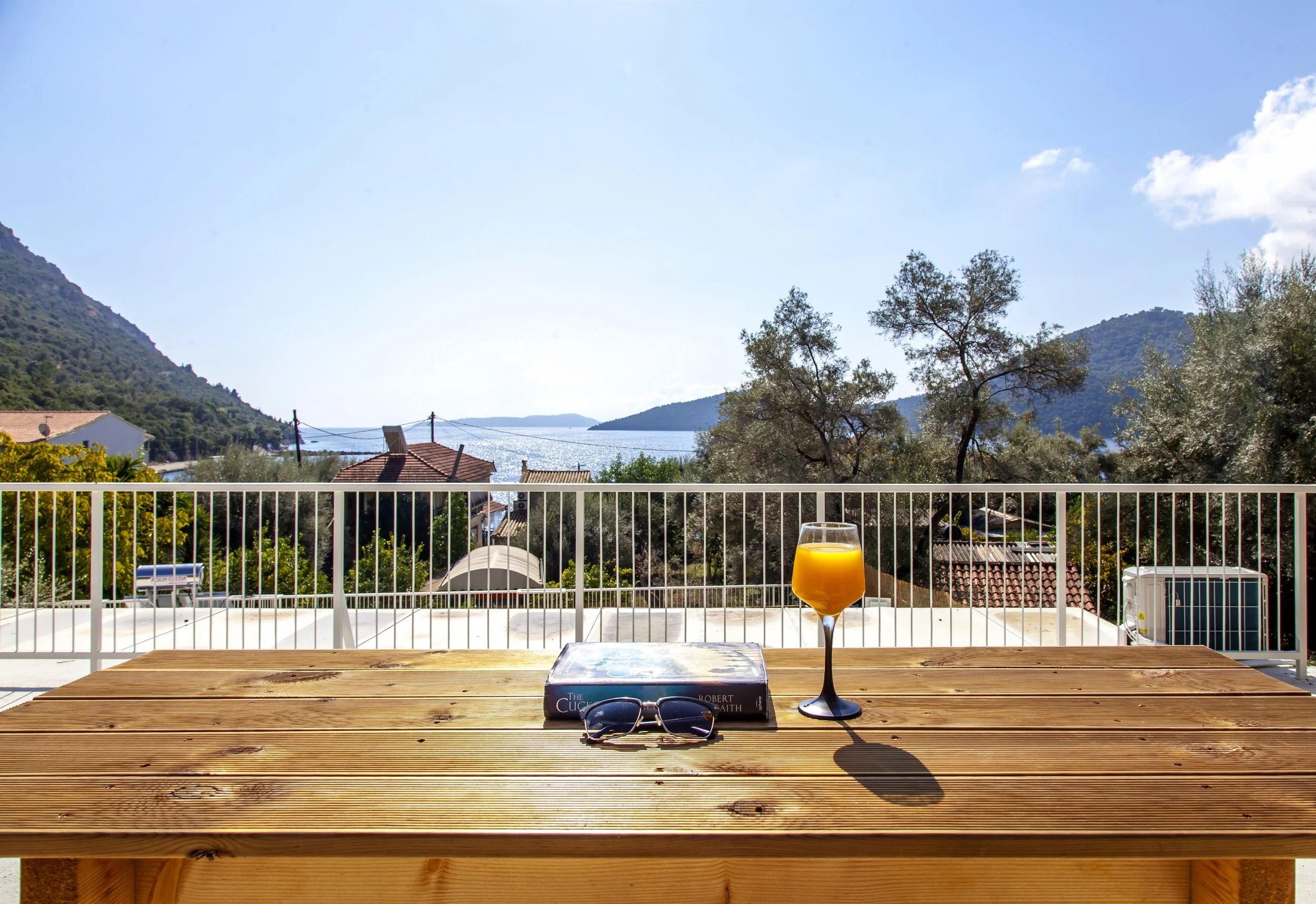 A view from a balcony with a wooden table, a glass of orange juice, a book, sunglasses, and a pair of keys. In the background, there are trees, houses with red-tiled roofs, hills, and a body of water under a clear blue sky with a few clouds.