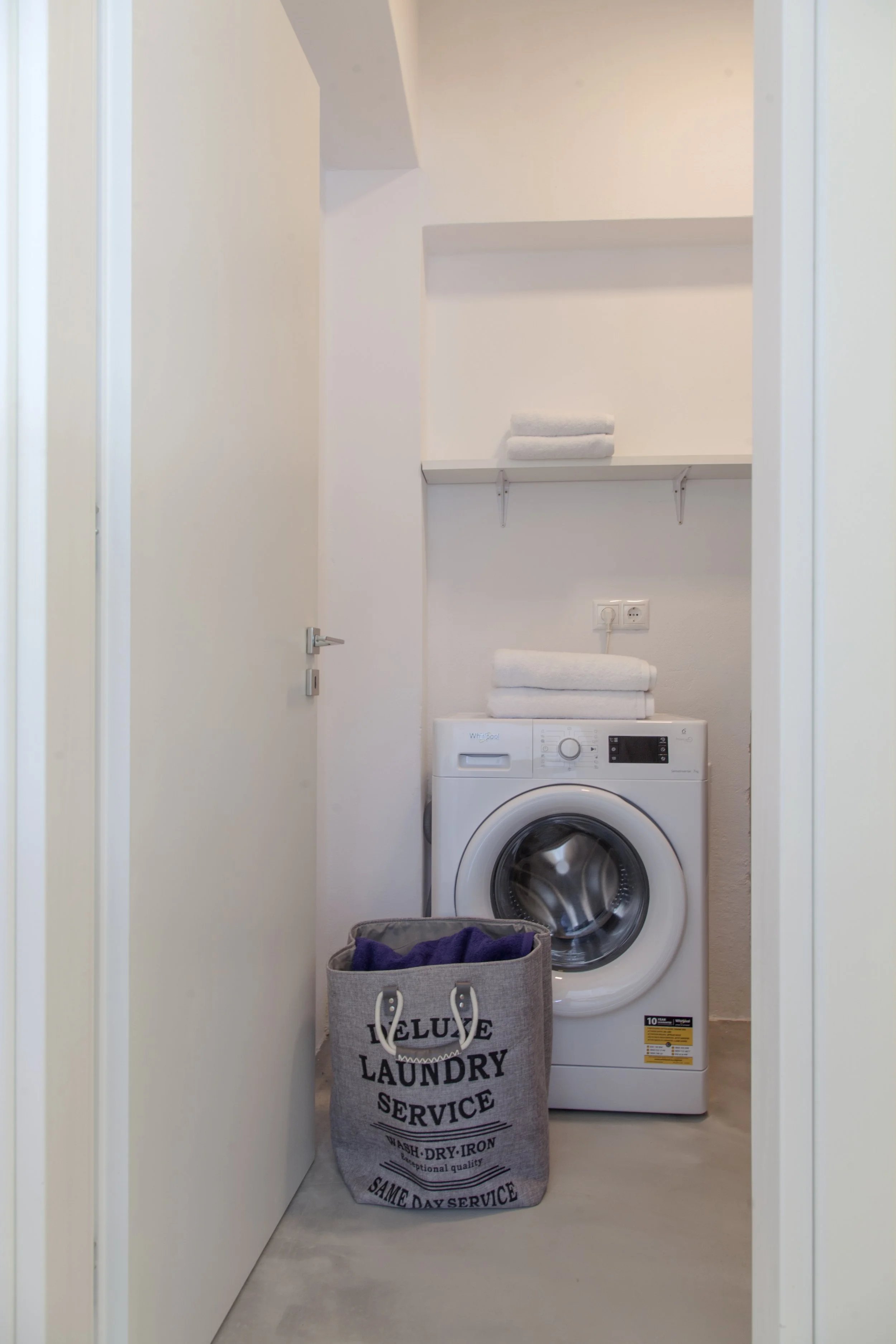 A laundry room with a front-loading washing machine, white shelves with folded towels, an electrical outlet, and a laundry basket with towels. A laundry bag with the words 'Luxury Laundry Service' is placed in front of the machine.