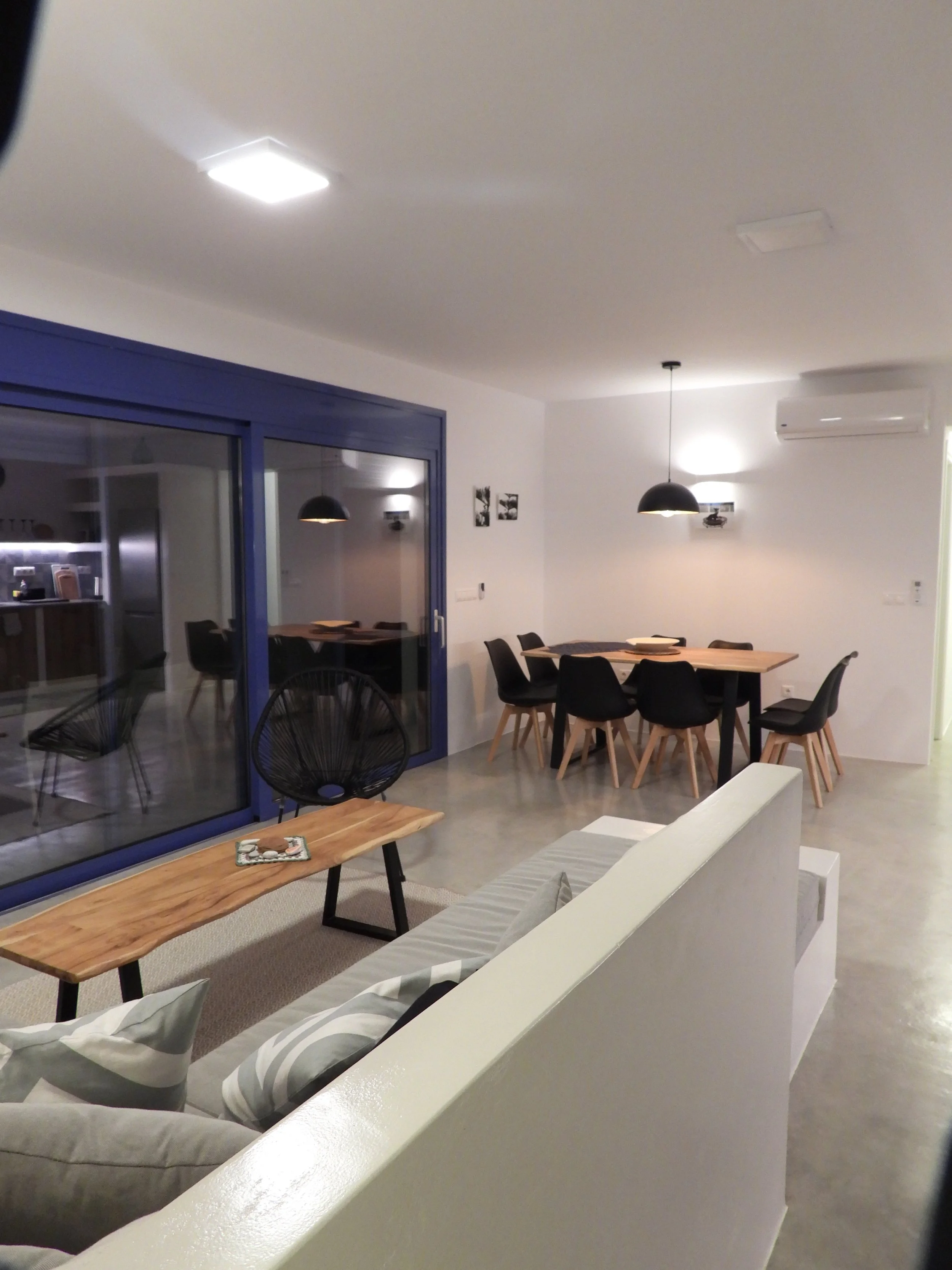 Living room and dining area in a modern home, with a white sofa with striped cushions, a wooden coffee table, black chairs around a wooden dining table, pendant and ceiling lights, and blue sliding glass doors.