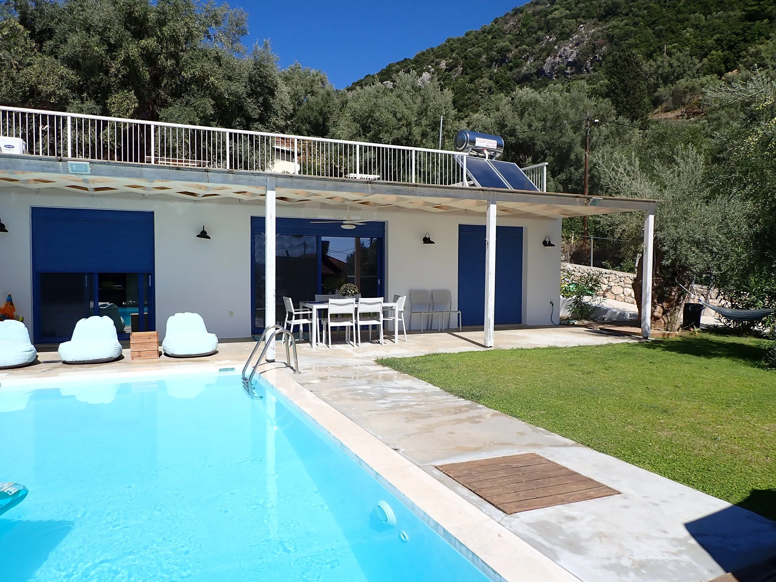 View of a backyard with a swimming pool, outdoor table and chairs, a building with blue doors, and a hillside covered with trees and shrubs in the background.