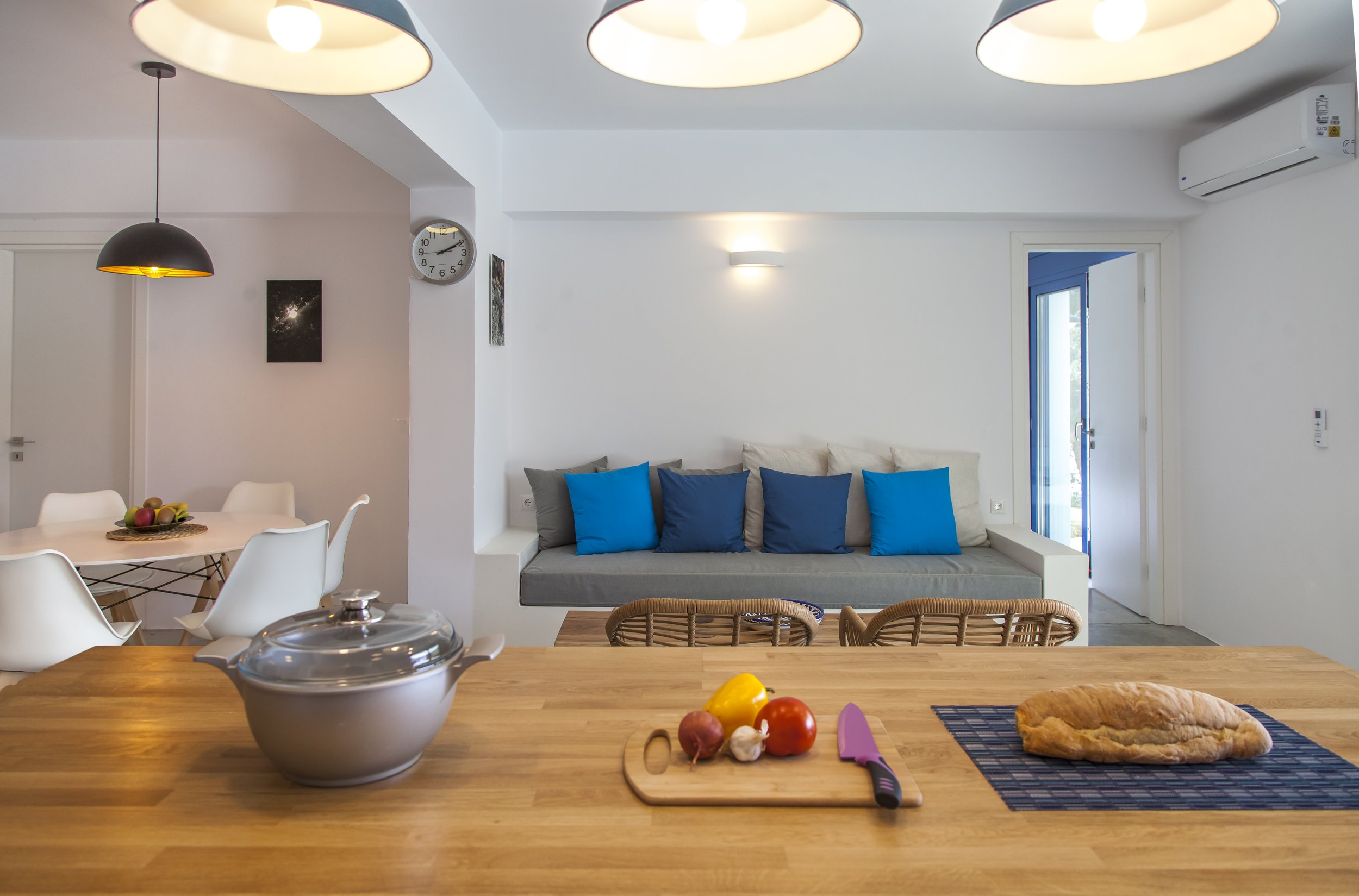 Kitchen with wooden countertop. Items on counter include a large metal pot, tomatoes, garlic, a yellow pepper, a pink spatula, and a small slice of pizza on a striped placemat. In the background, there is a dining area with white chairs and a dining table. The living area has a white sofa with blue and gray cushions, a wall clock, and wall art. The ceiling has large round light fixtures, and there is a window with a blue frame. An air conditioning unit is mounted on the wall.