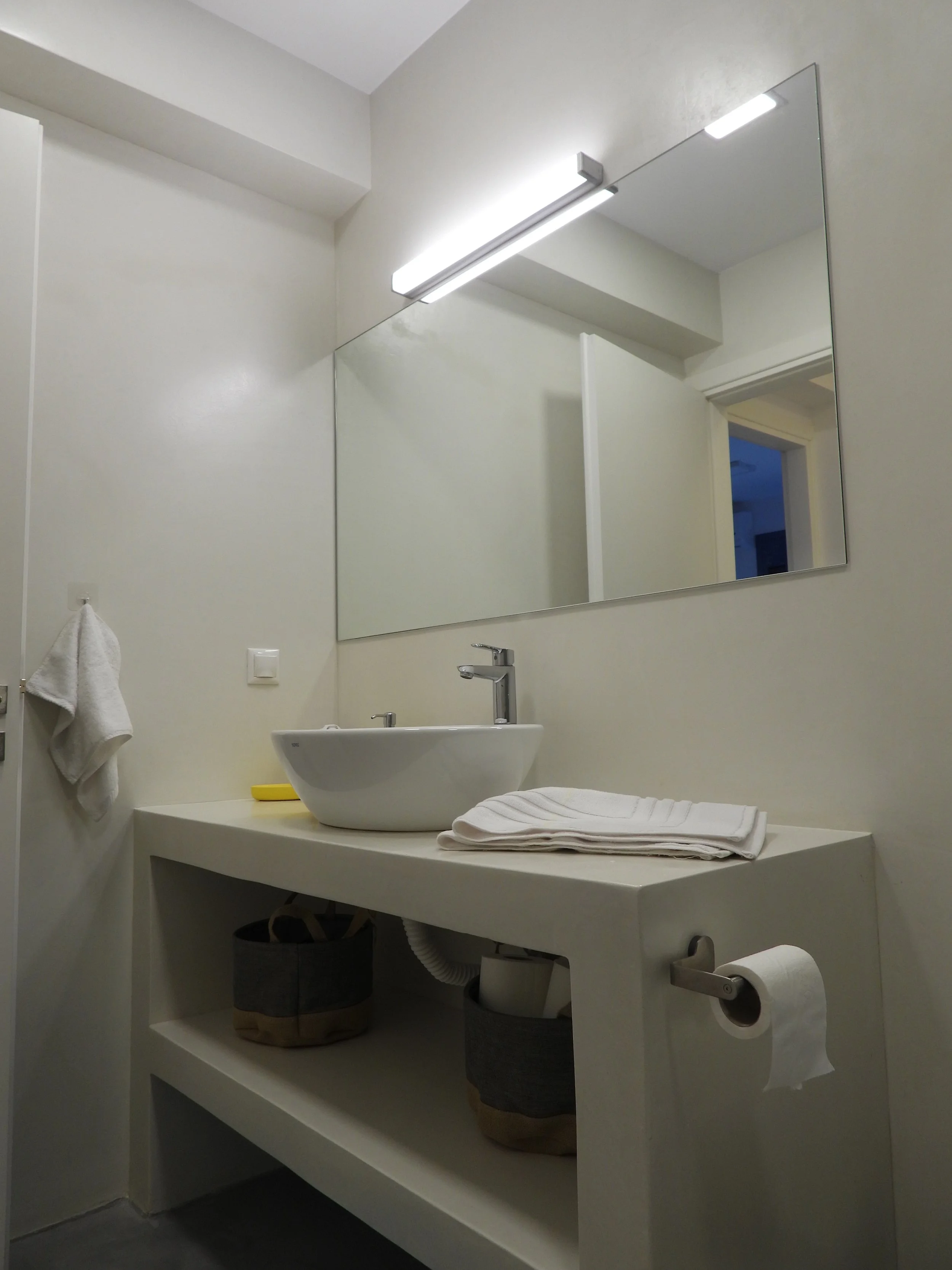 A minimalist bathroom with a wall-mounted mirror, a small white sink with a modern faucet, a shelf holding folded white towels and storage baskets, a towel hanging on a hook, and a toilet paper roll on a holder attached to the side of the shelf.