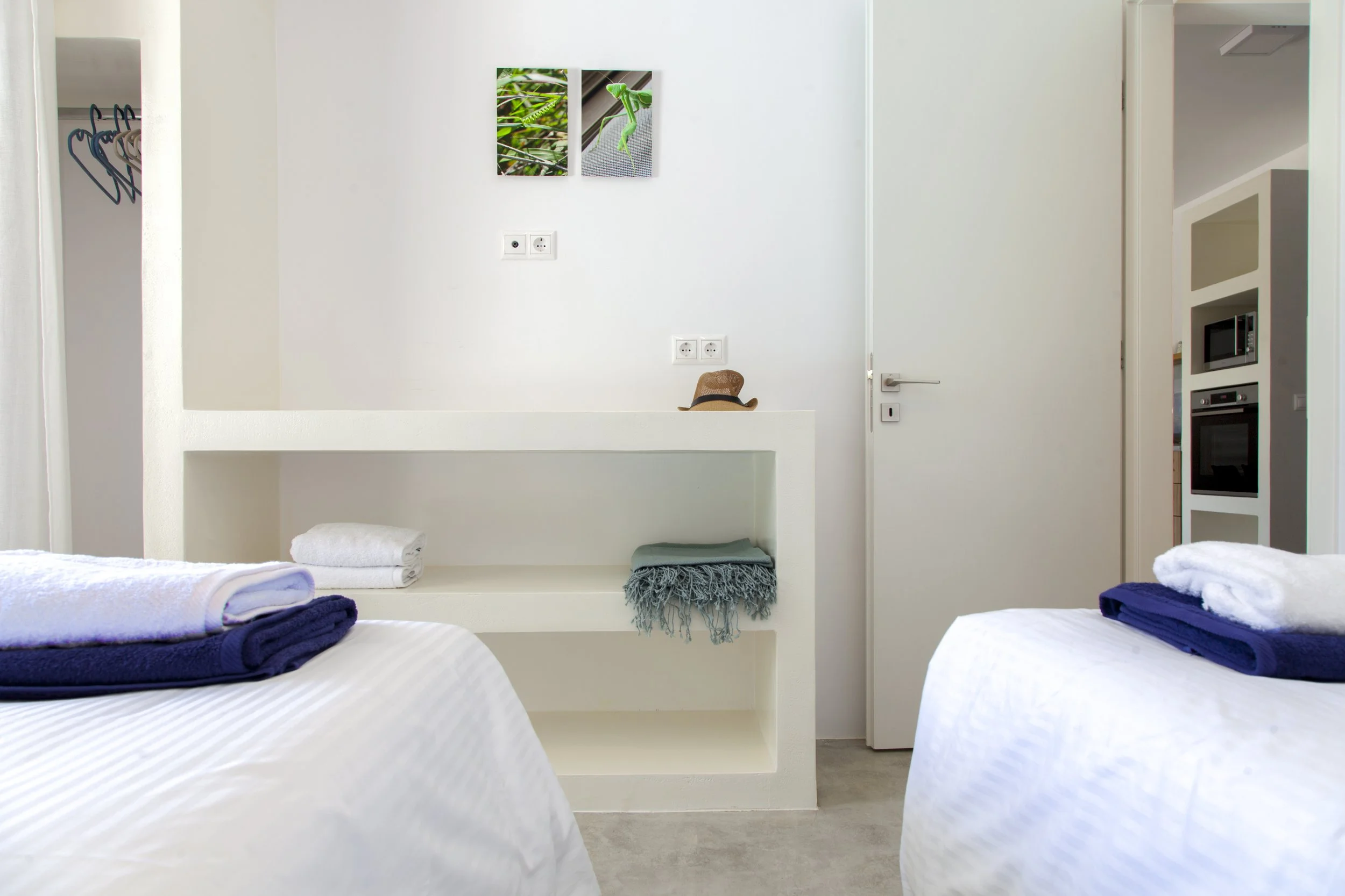 A hotel room with two beds each with folded towels, a middle shelf with a hat and towels, and a white wall with two photographs of green leaves and a chameleon, and electrical outlets.