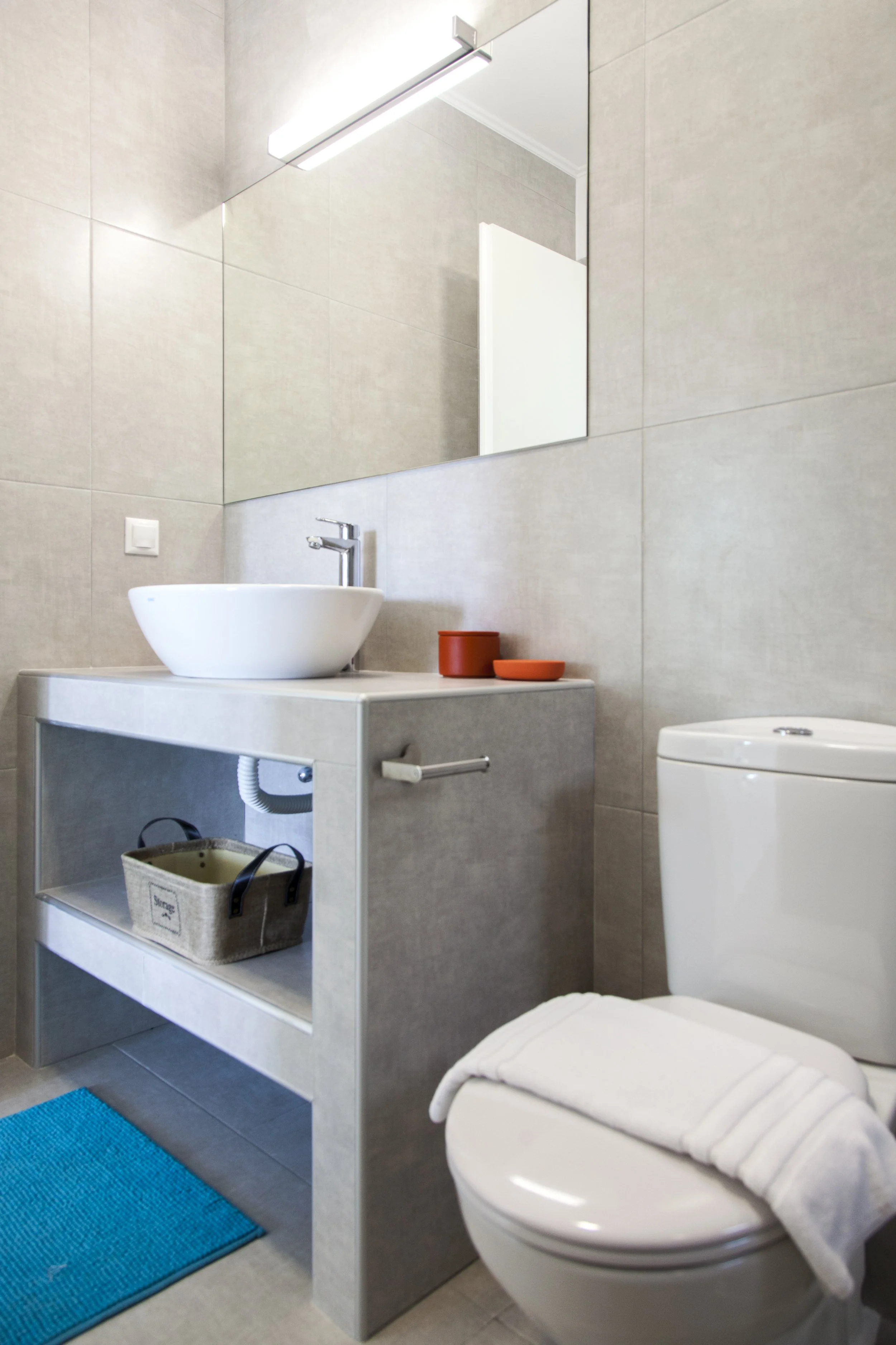 A modern bathroom with beige tiled walls, a large rectangular mirror, and a white vessel sink on a light-colored vanity. There is a blue bath mat on the floor and a white toilet with a towel draped over the lid. A small wicker basket sits inside the 