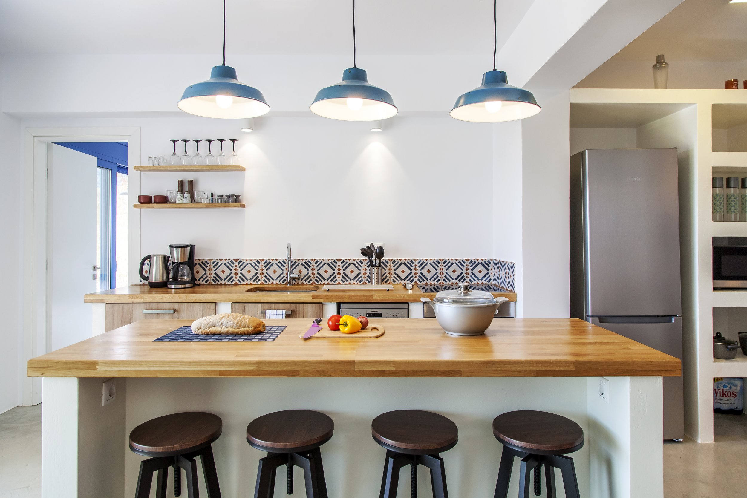 Modern kitchen with wooden island counter, blue pendant lights, patterned tile backsplash, stainless steel refrigerator, open shelving, and barstools.