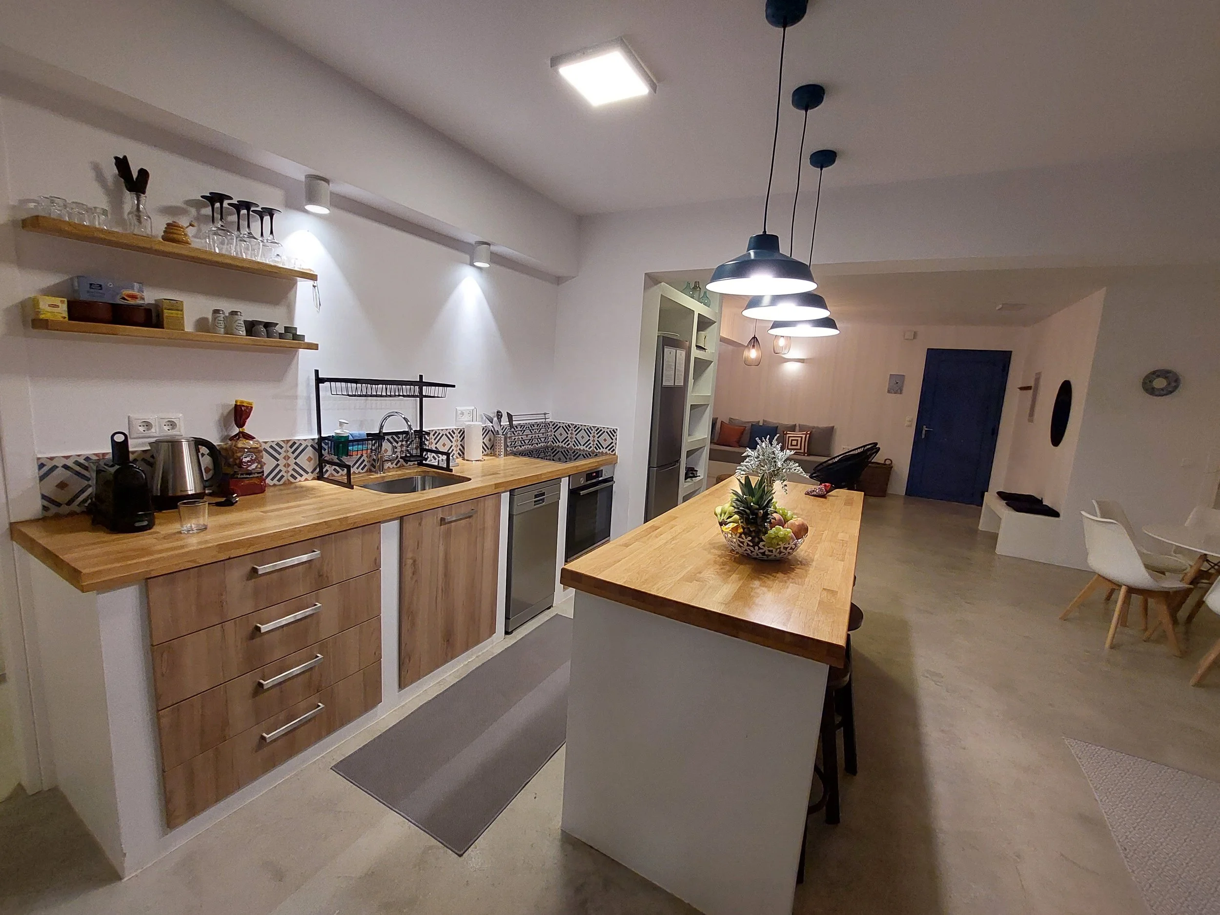 Open-concept kitchen with wooden countertops, white cabinetry, a stainless-steel oven and stovetop, black and white geometric tile backsplash, hanging pendant lights, a white shelving unit, and a dining area with chairs and a floral centerpiece.