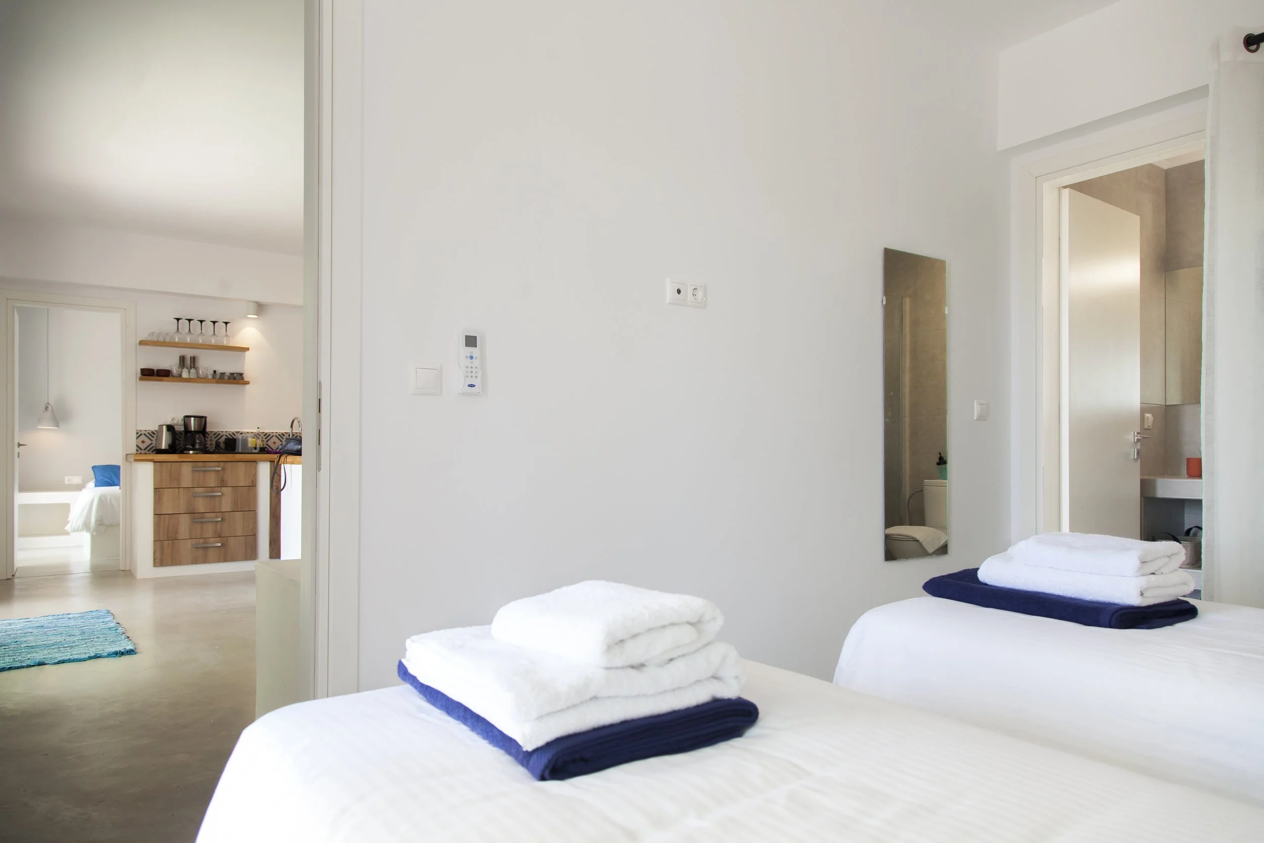 A bright bedroom with two beds covered in white linens and folded towels, a mirrored wardrobe, and a view into a small kitchen area with shelves and appliances.