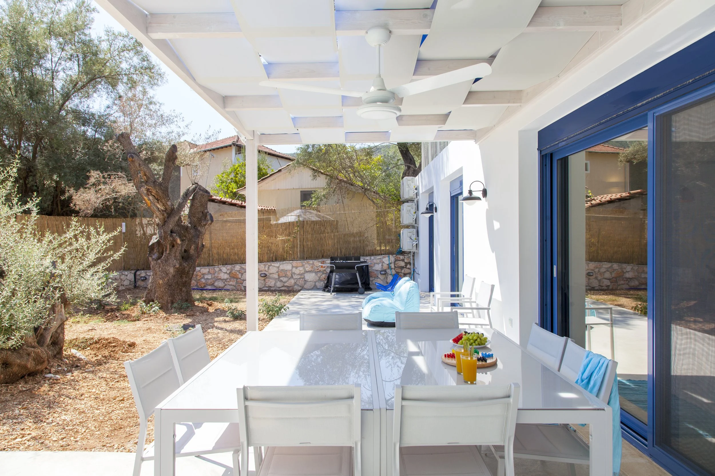 Outdoor patio with a white dining table, chairs, and a bowl of fruit, next to a house with blue sliding doors and a white ceiling fan, surrounded by trees and neighboring houses.