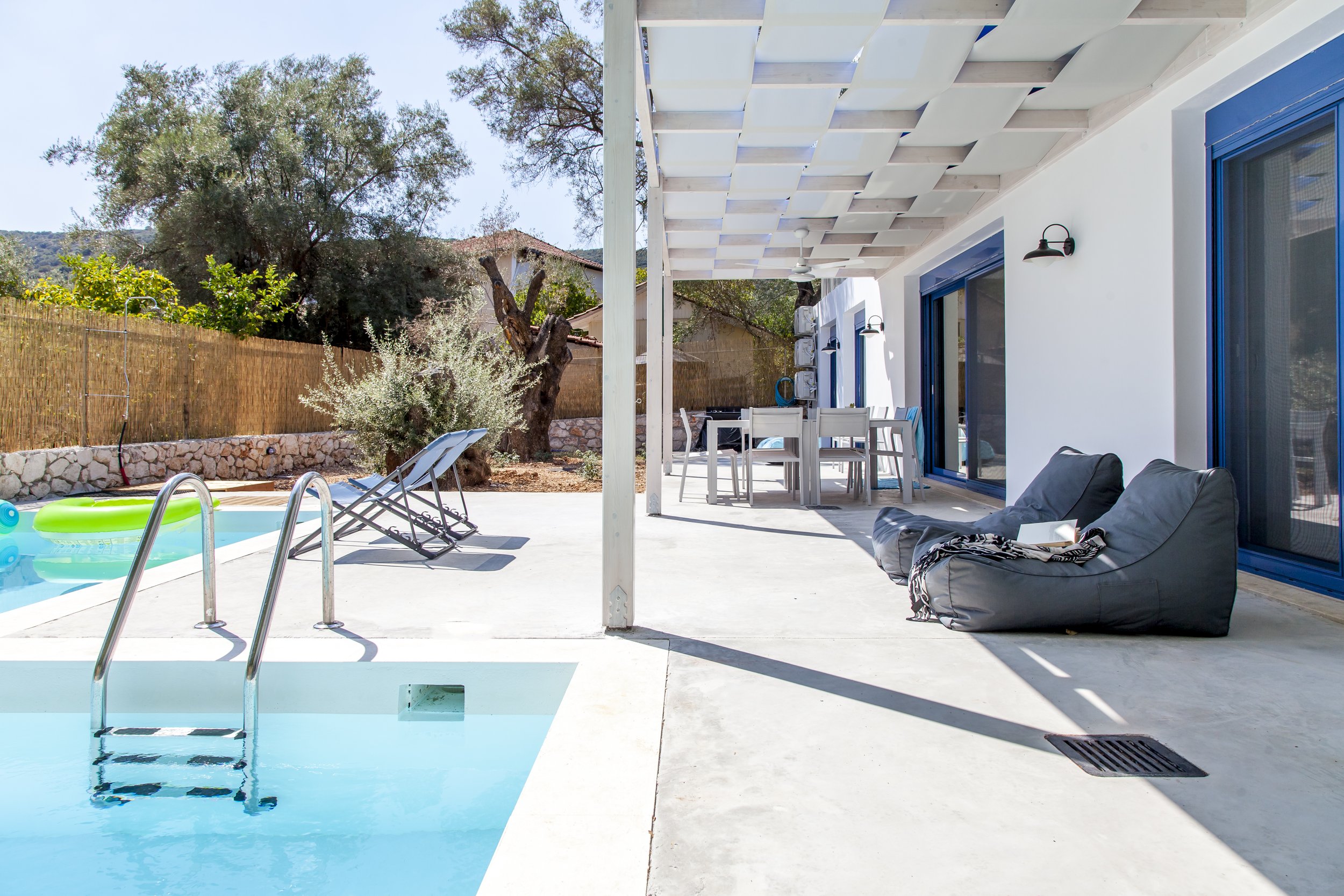 Backyard pool area with blue water, white concrete deck, pool ladder, lounge chairs, outdoor dining furniture, large tree, and a white house with blue window frames and sliding doors.