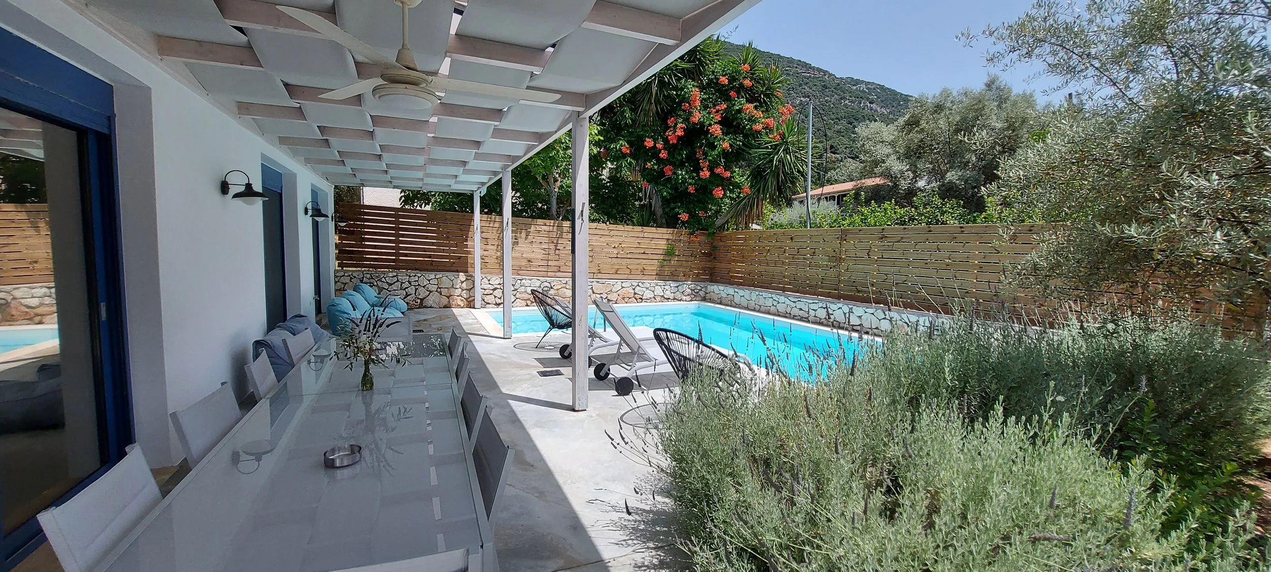 Outdoor patio with a dining table, chairs, lounge chairs, a swimming pool, and a wooden fence, surrounded by green trees and a mountainous landscape.