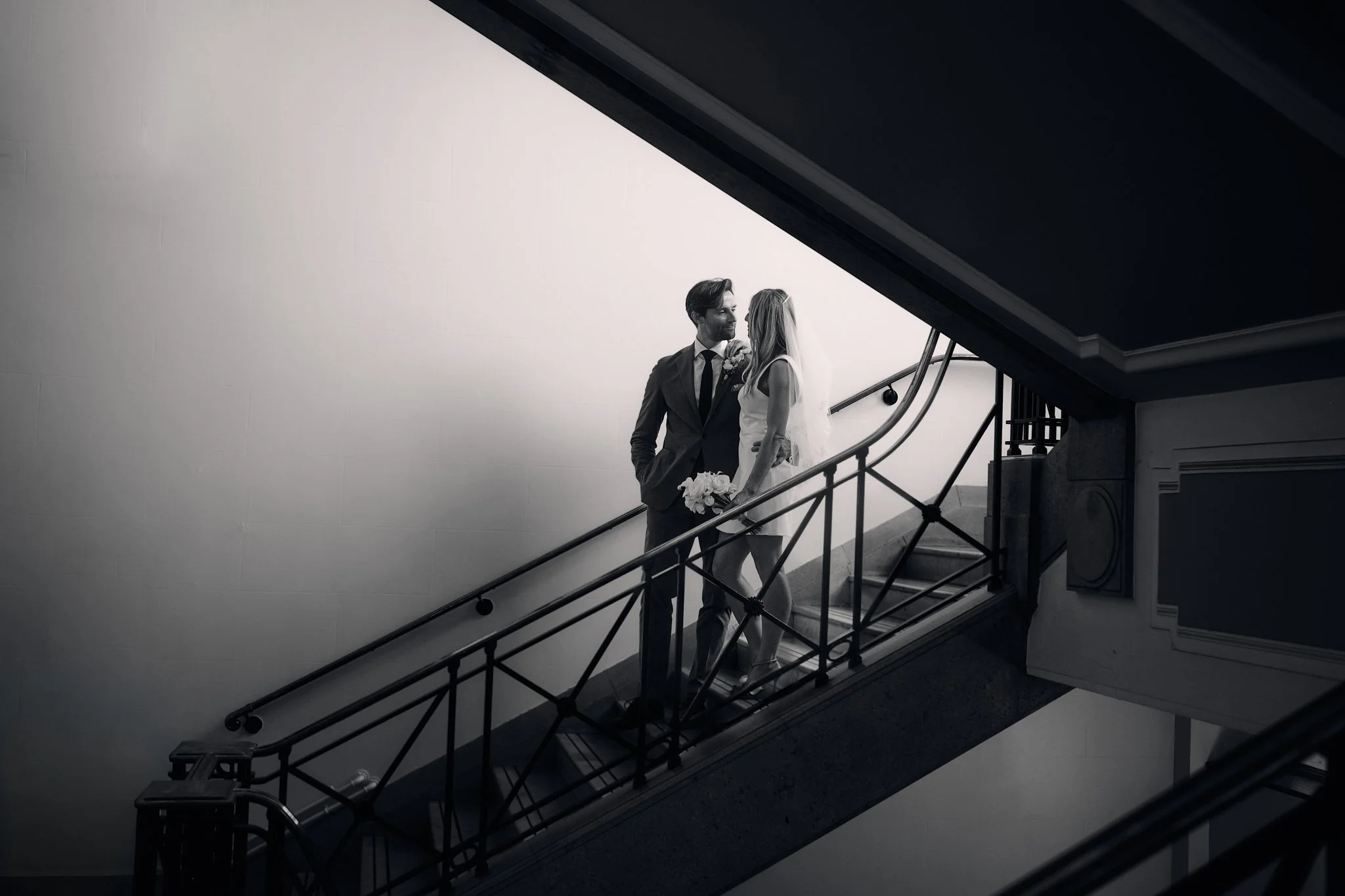 Black and white photo of a couple in wedding attire standing on a staircase, facing each other and smiling in an indoor setting.