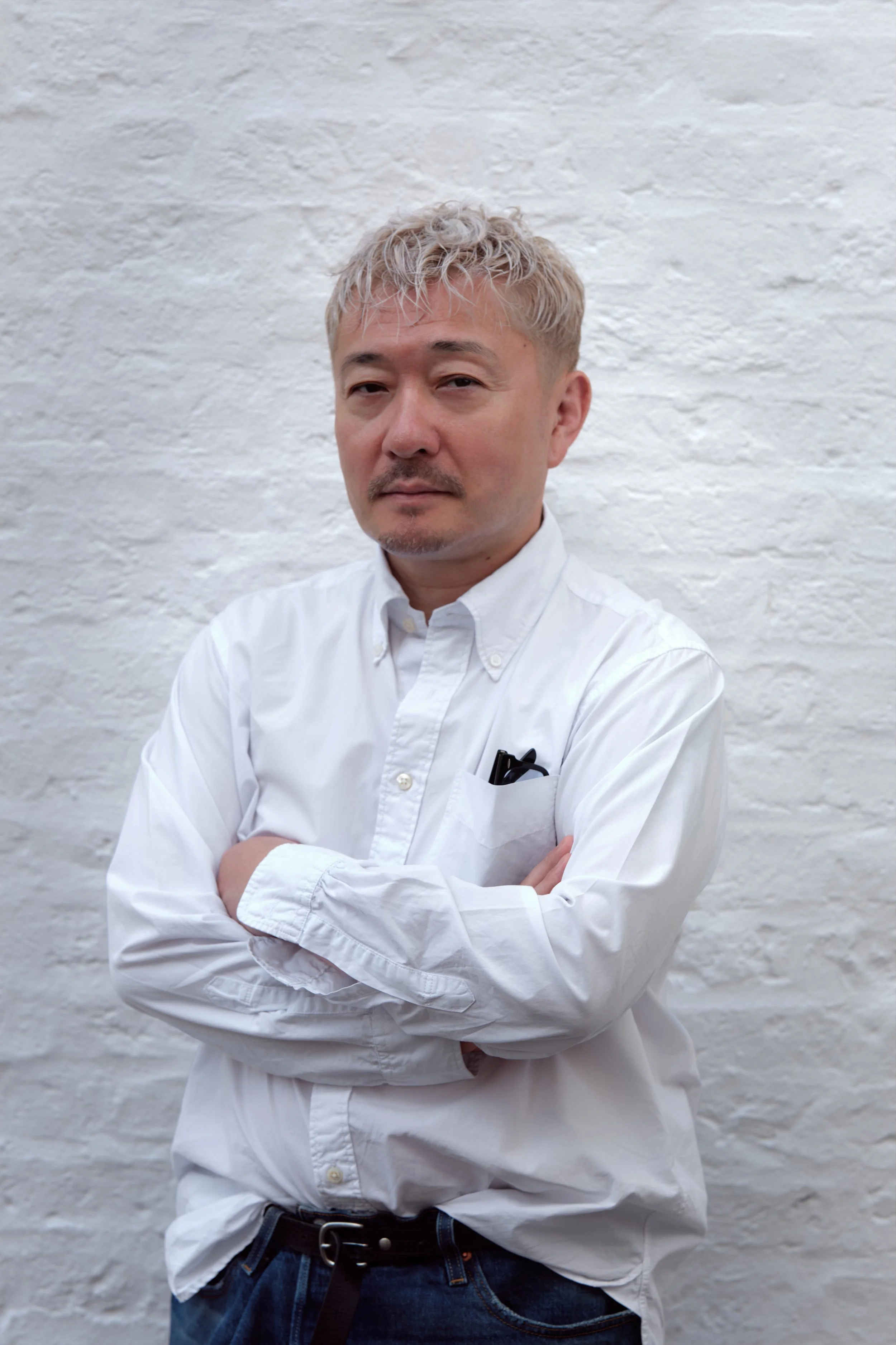 A man with short, curly, blonde hair, wearing a white button-up shirt with the sleeves rolled up, standing with arms crossed in front of a white textured wall.
