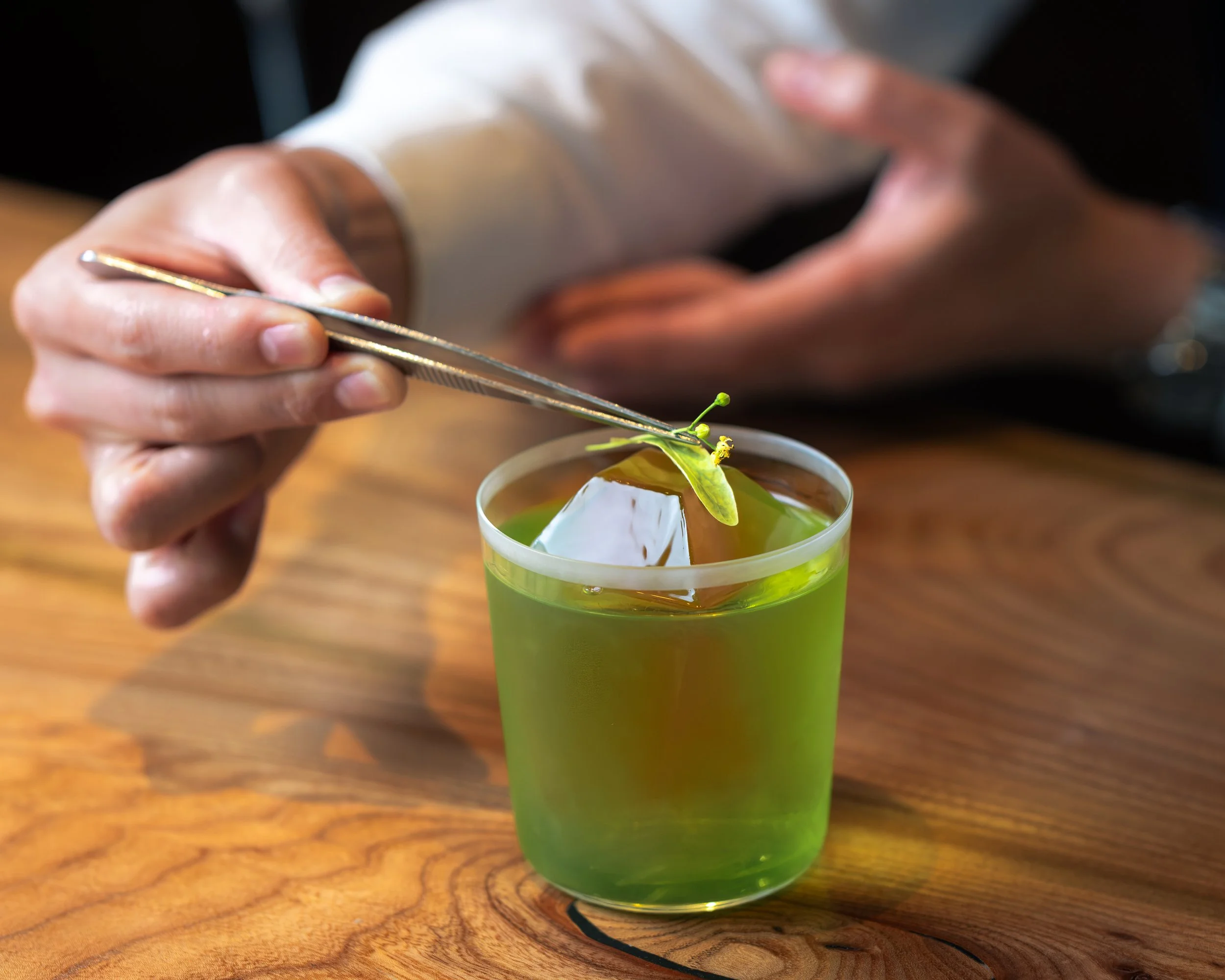 A hand holding chopsticks picking a small flower from a glass of green-colored beverage on a wooden table.