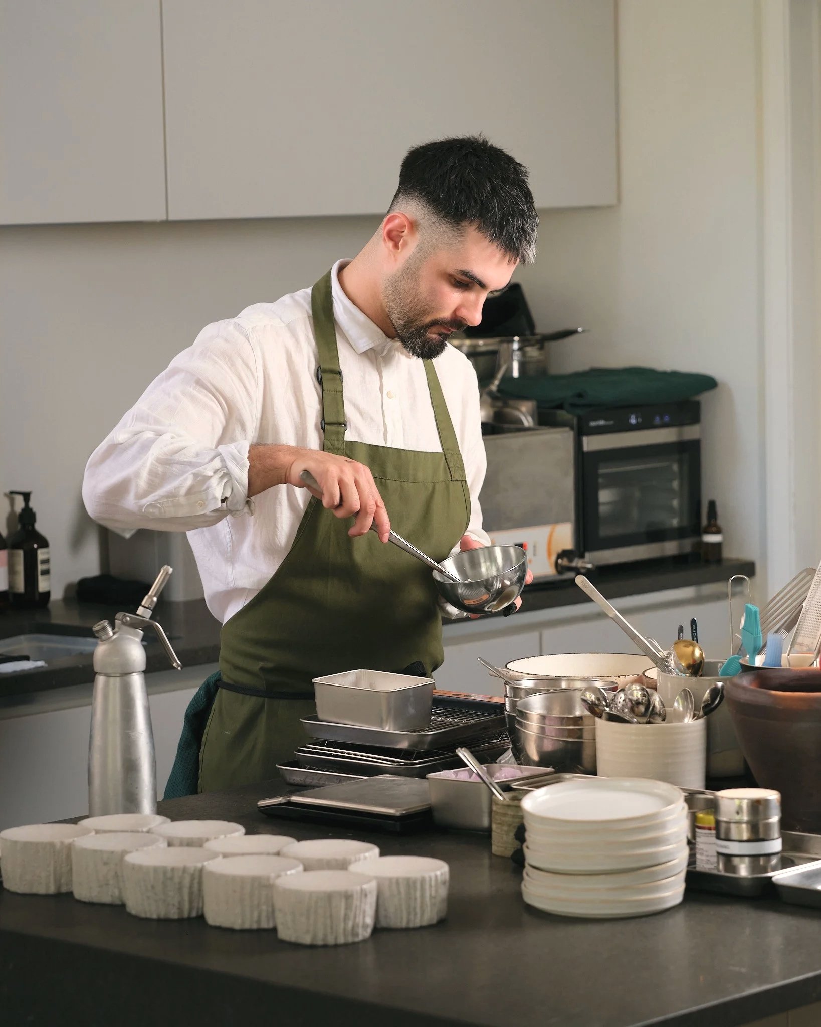 A man in a white shirt and green apron preparing food in a commercial kitchen. The countertop has various dishes and kitchen tools, with stacked white bowls and a metallic container in the foreground.