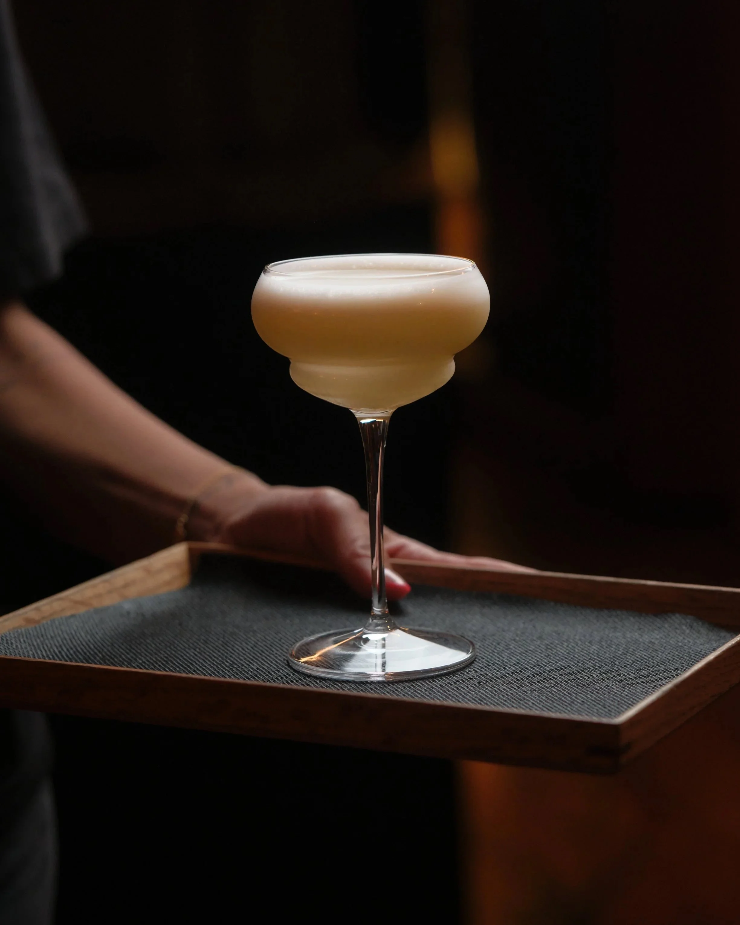 Elegant cocktail in a coupe glass with a frothy top on a wooden tray, hand with red nail polish rests nearby.