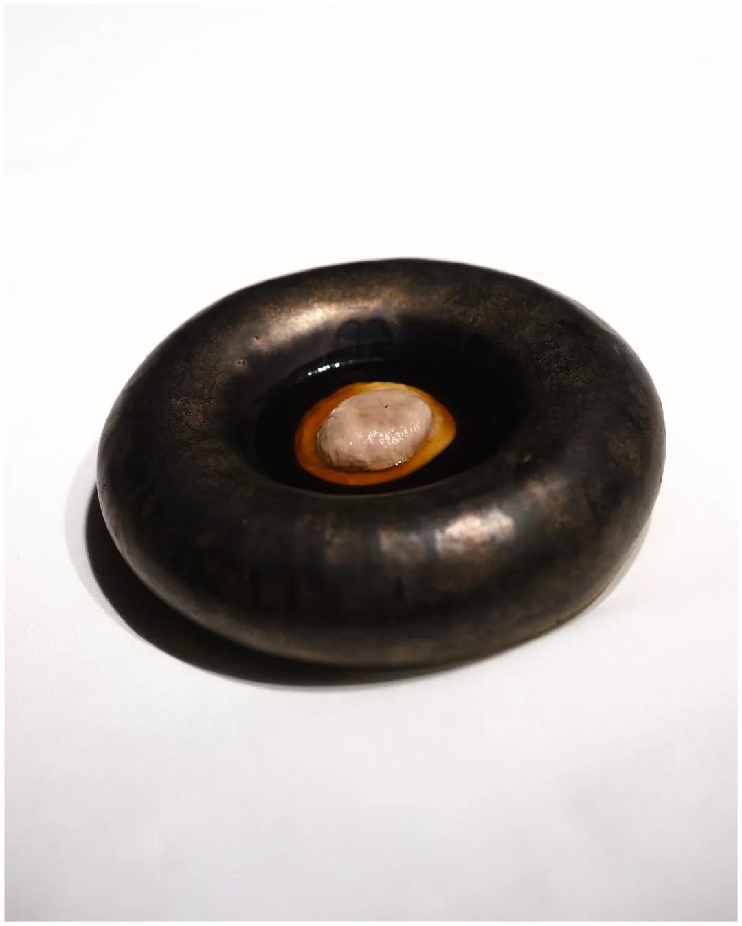 A small, round, black stone with a hollowed-out center, containing a beige and white object, resembling a shell or a worm, against a white background.