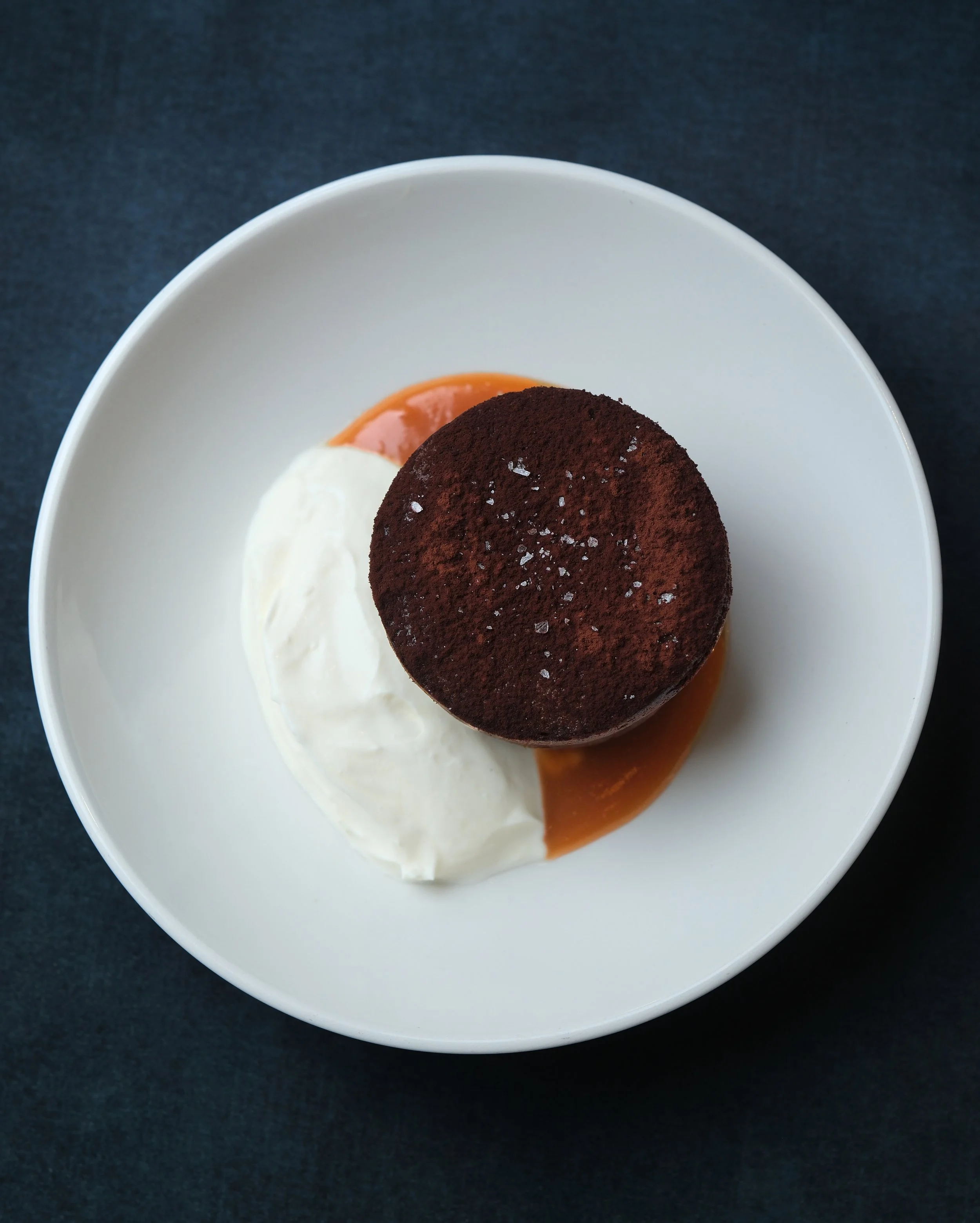 Close-up of a dessert with a dark brown cocoa powder topping on a round cake or pastry, served with whipped cream and caramel sauce on a white plate, against a dark background.