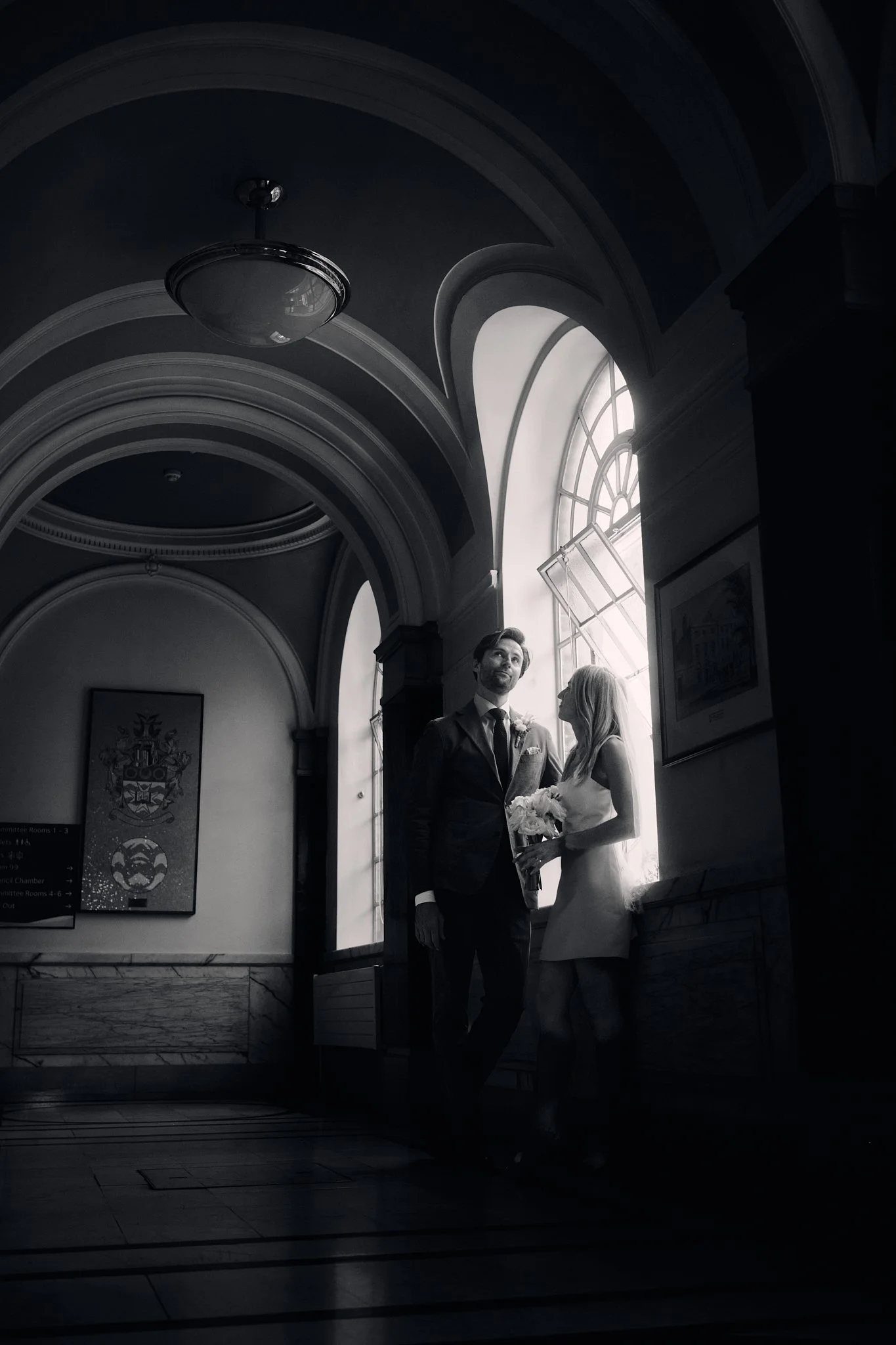 A black and white photo of a bride and groom standing close together in a building with an arched ceiling and large window. The bride holds a bouquet and the groom looks thoughtfully into the distance.