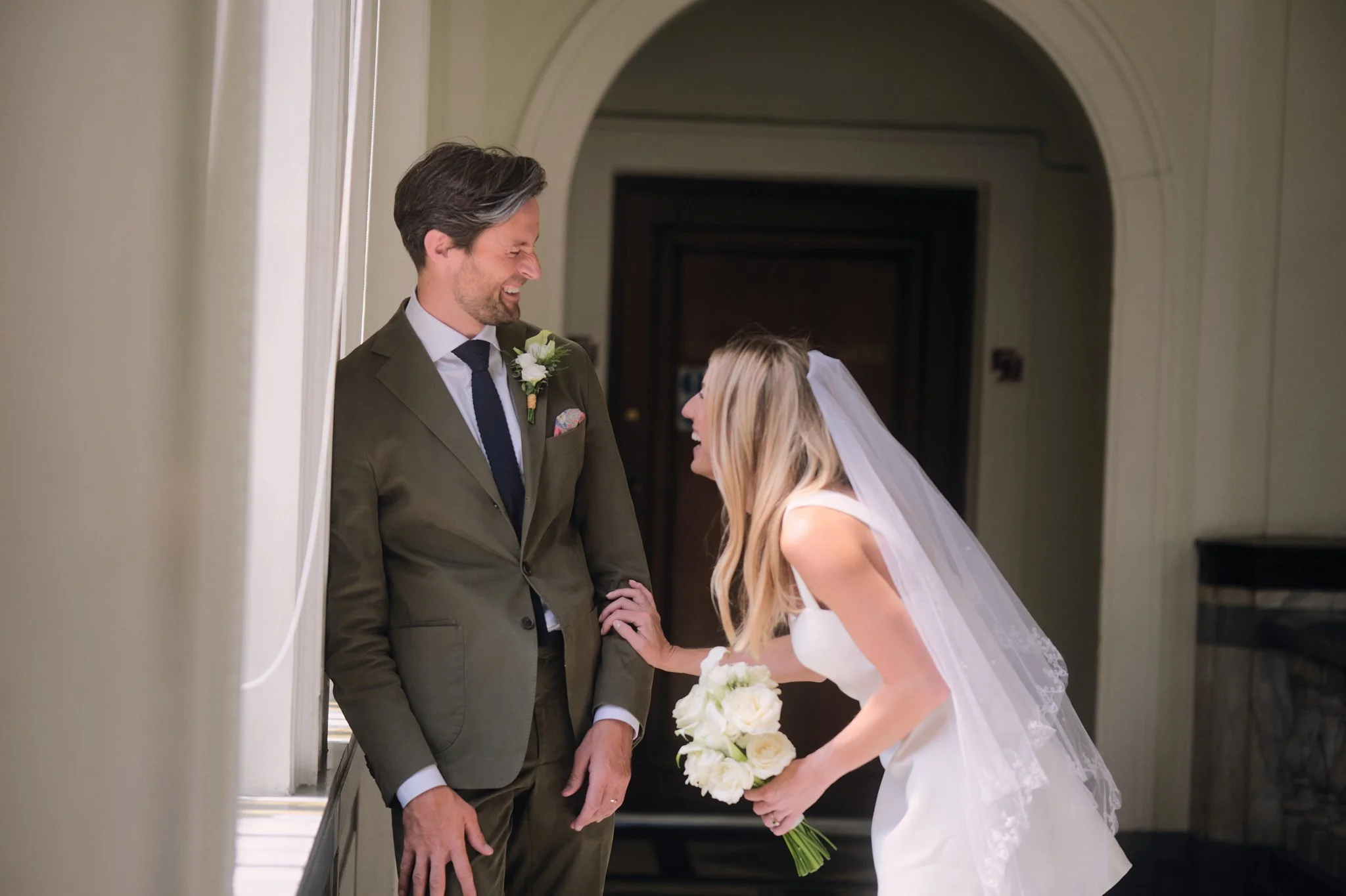 A bride and groom sharing a joyful moment during their wedding, standing in a hallway; the bride is holding a bouquet of white roses and the groom is wearing a suit with a boutonniere, both smiling and looking at each other.