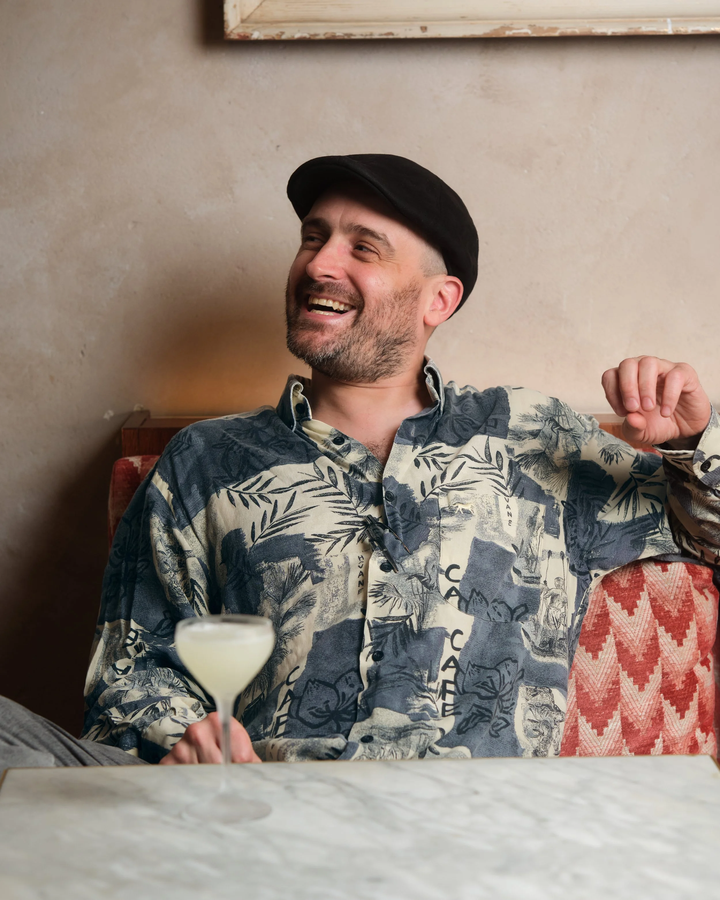 A man smiling and enjoying a drink at a restaurant, holding a cocktail glass with a pale drink in front of him, wearing a black hat and a patterned shirt, seated at a table with a marble top, in a cozy, warmly lit setting.