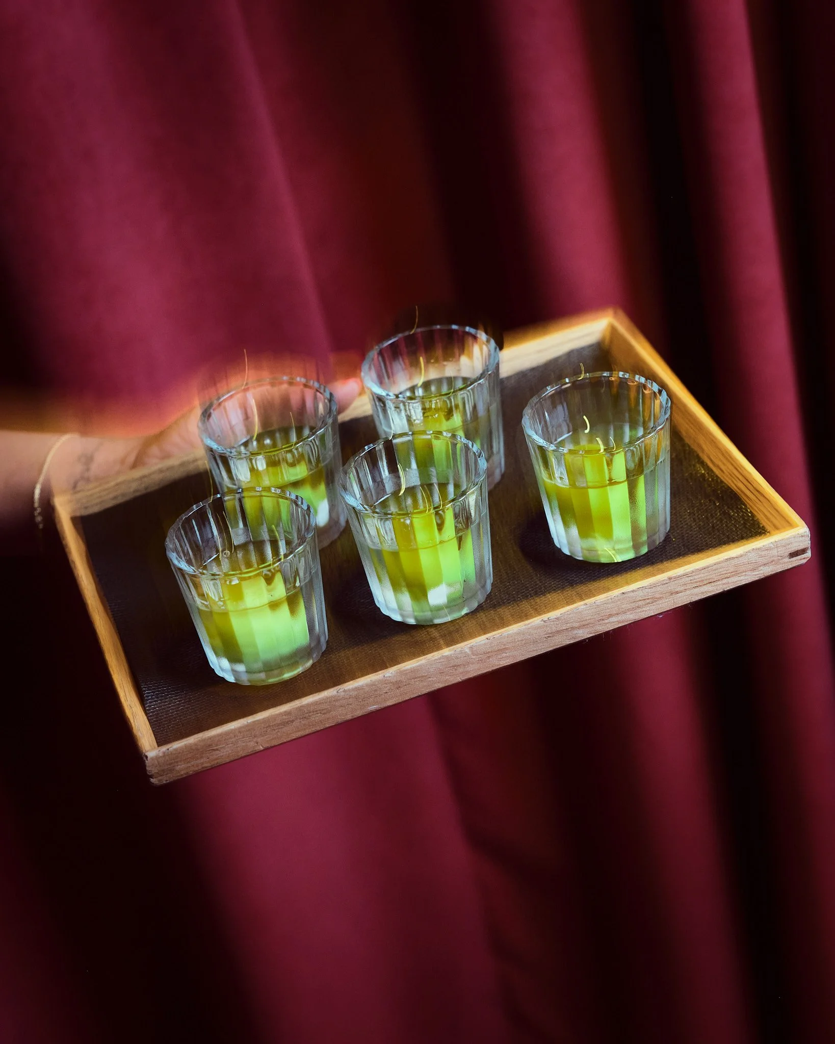 A wooden tray holding several small glasses filled with a yellow-green liquid, with a red velvet curtain in the background.