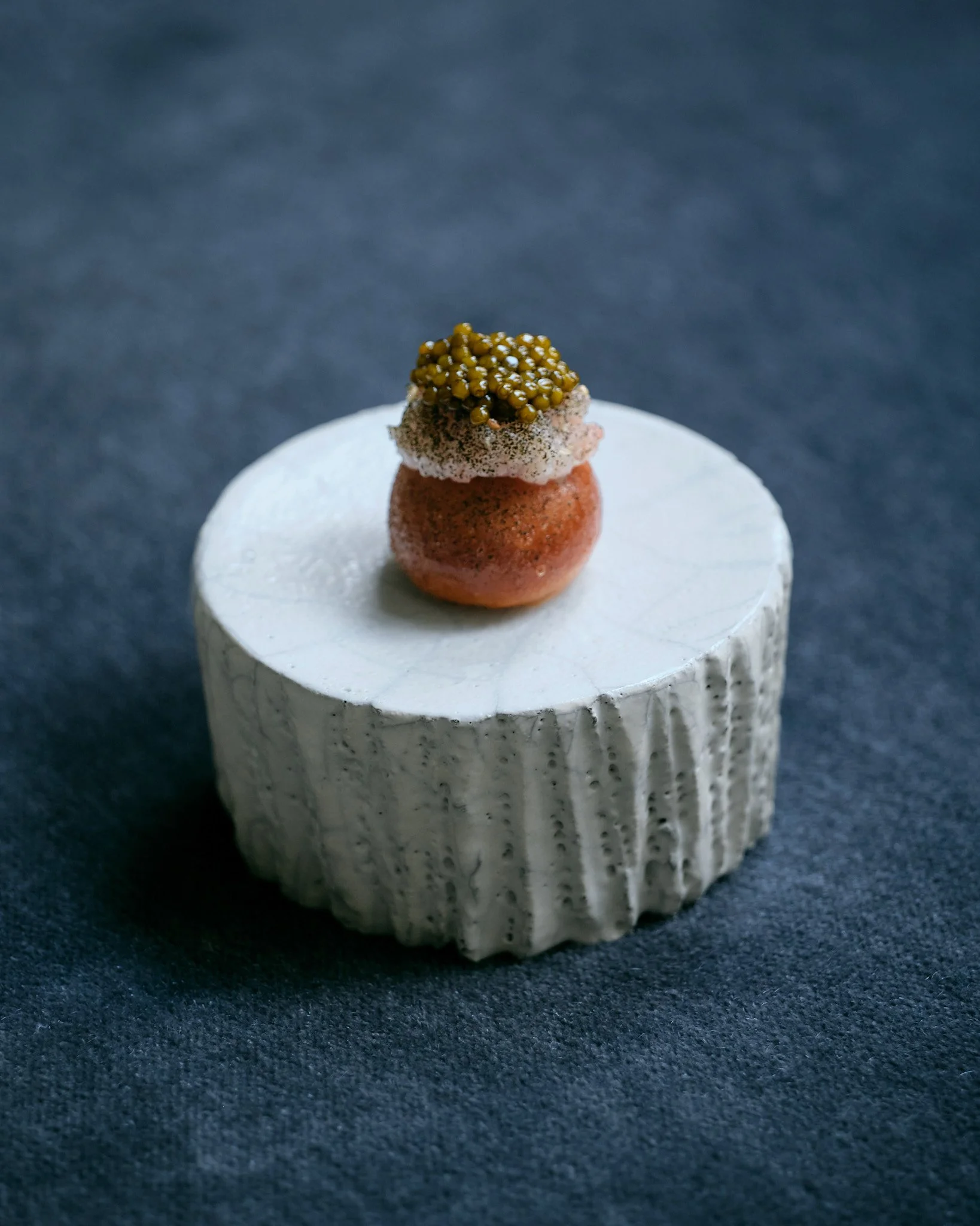 A small, layered pastry with a light pink layer, topped with a dollop of cream and yellowberries, placed on a white textured plate against a dark background.