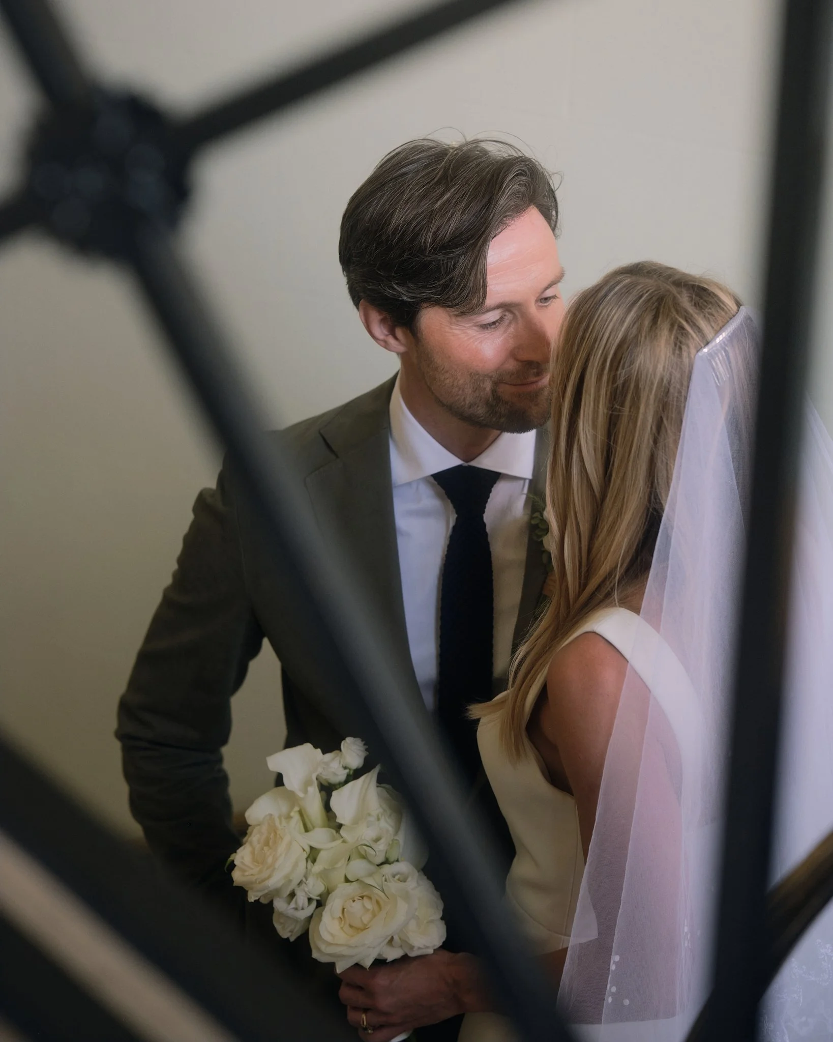 A groom with dark hair and a beard in a gray suit holds a bouquet of white roses while embracing a bride with blonde hair in a white wedding dress during a wedding ceremony, seen through a blurred frame.