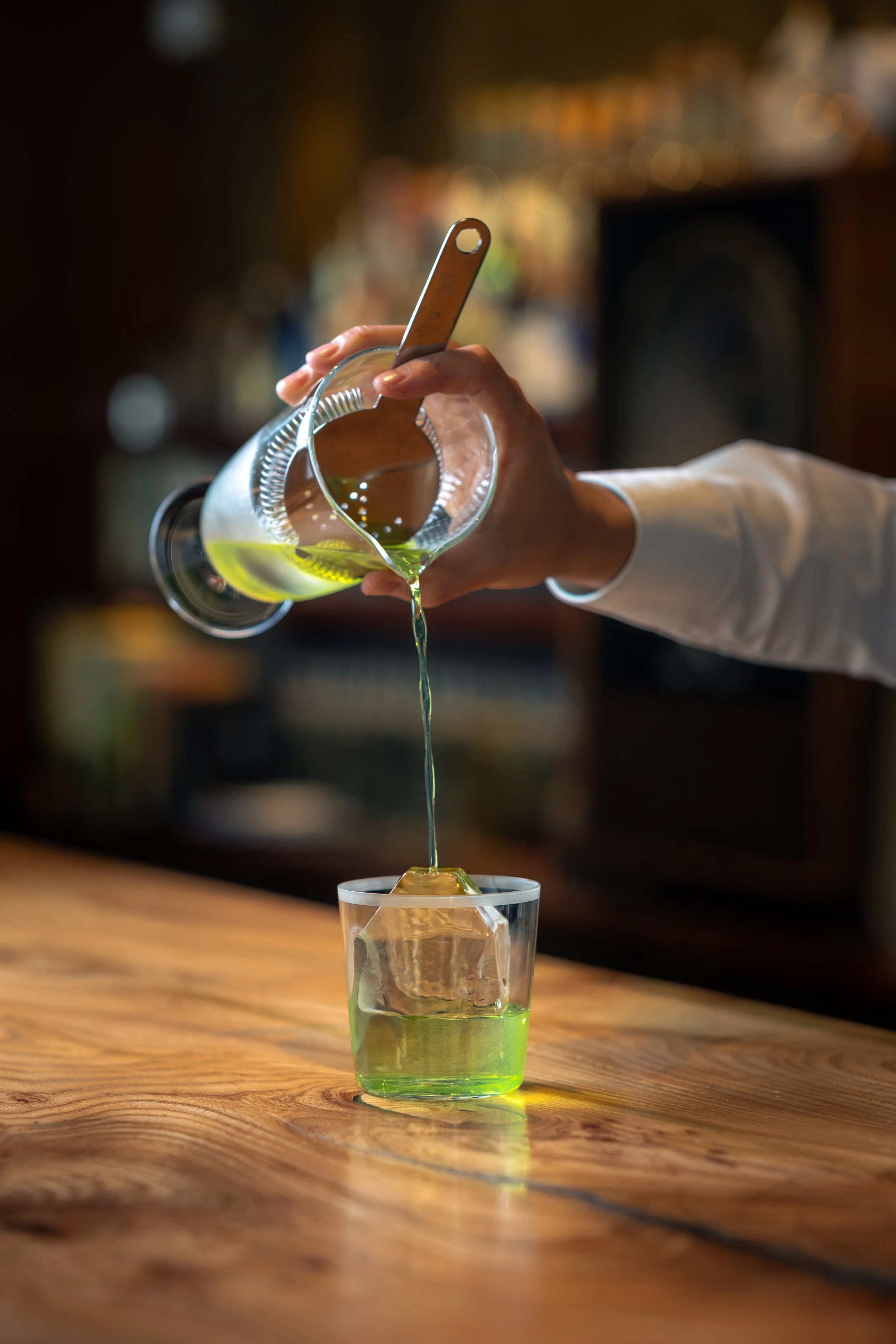 A hand is pouring green liquid from a glass pitcher into a glass on a wooden table.