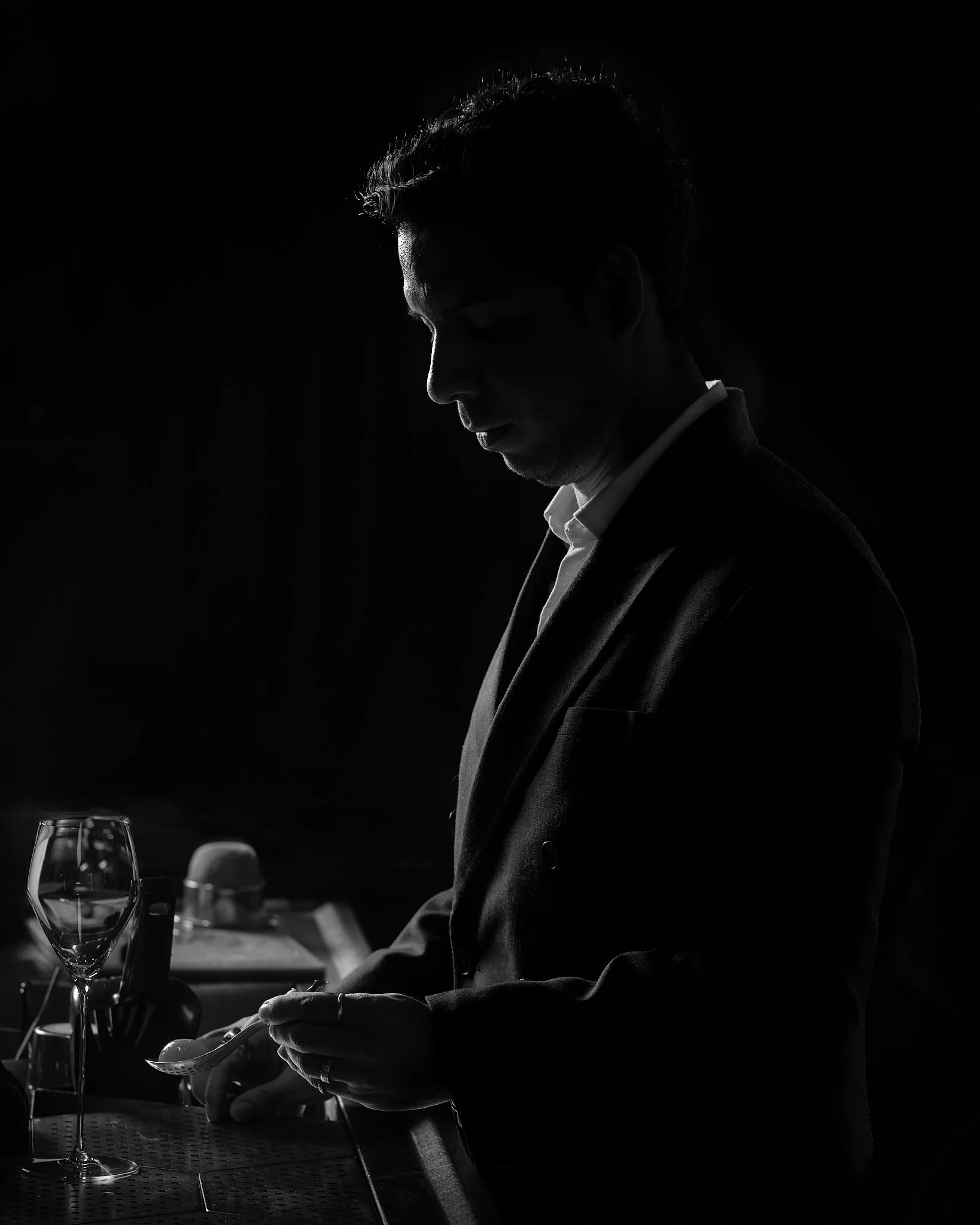 Side profile of a man in a suit, sitting at a table with a glass of wine and a small dish, in a dark, low-lit setting, appearing contemplative or focused.