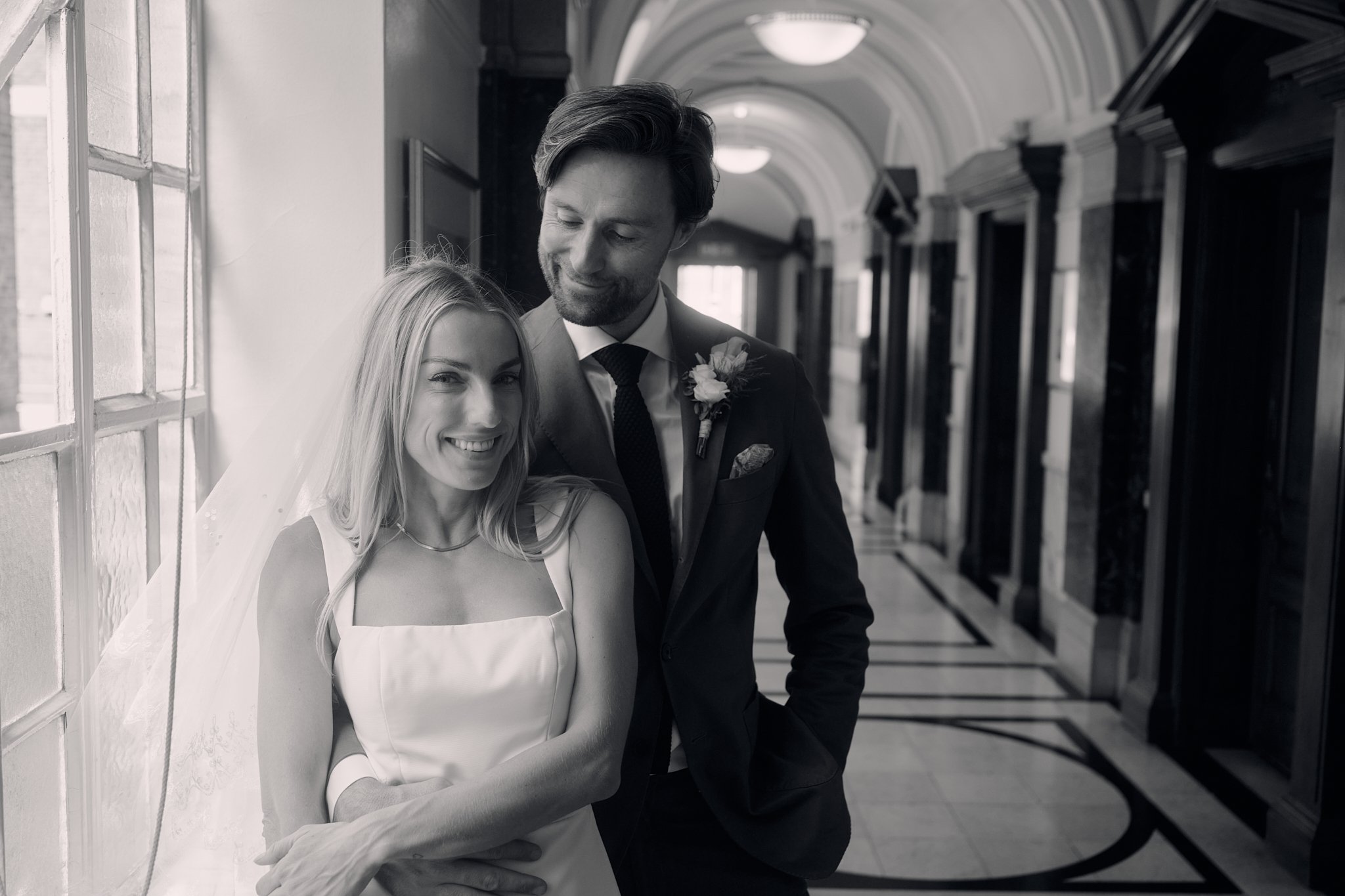 A black and white photo of a bride and groom laughing and embracing in an arched hallway with windows.