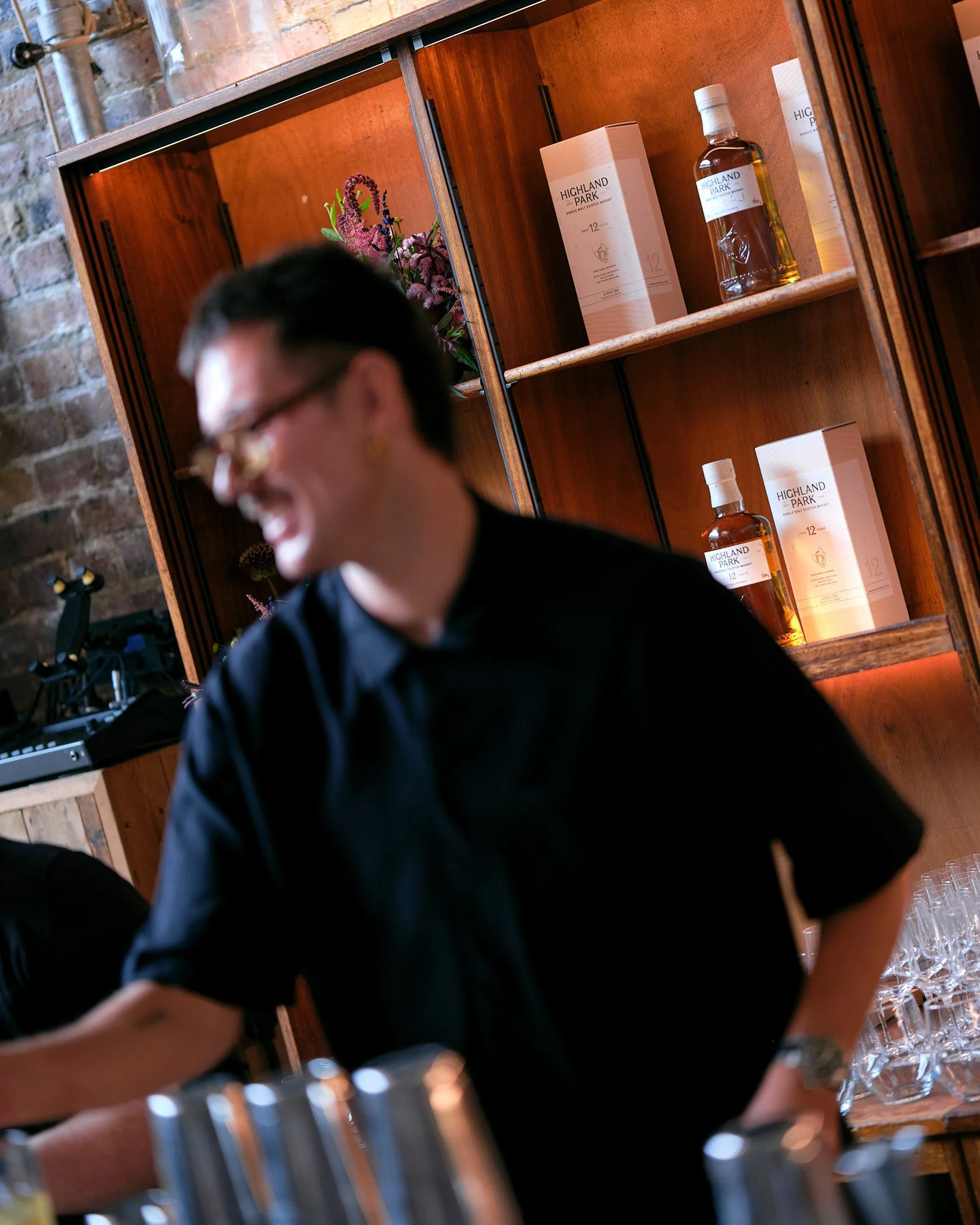 A blurry person with dark hair wearing glasses and a black shirt, seen from the side, with a wooden bookshelf behind them holding bottles of Highland Park single malt Scotch whisky and a floral decoration.