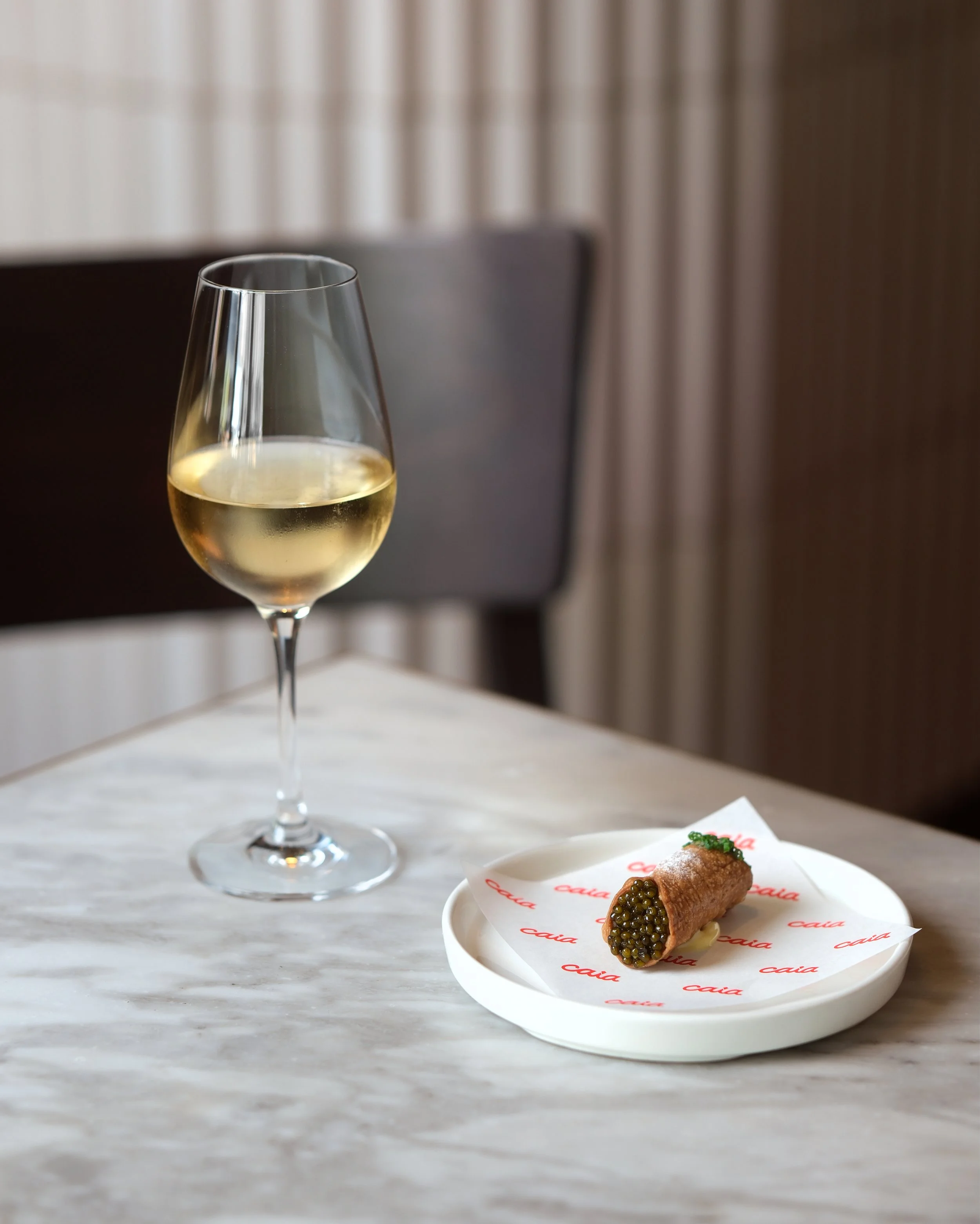 A glass of white wine and a cannoli pastry on a white plate with red branding, placed on a marble surface in a setting with beige curtains.