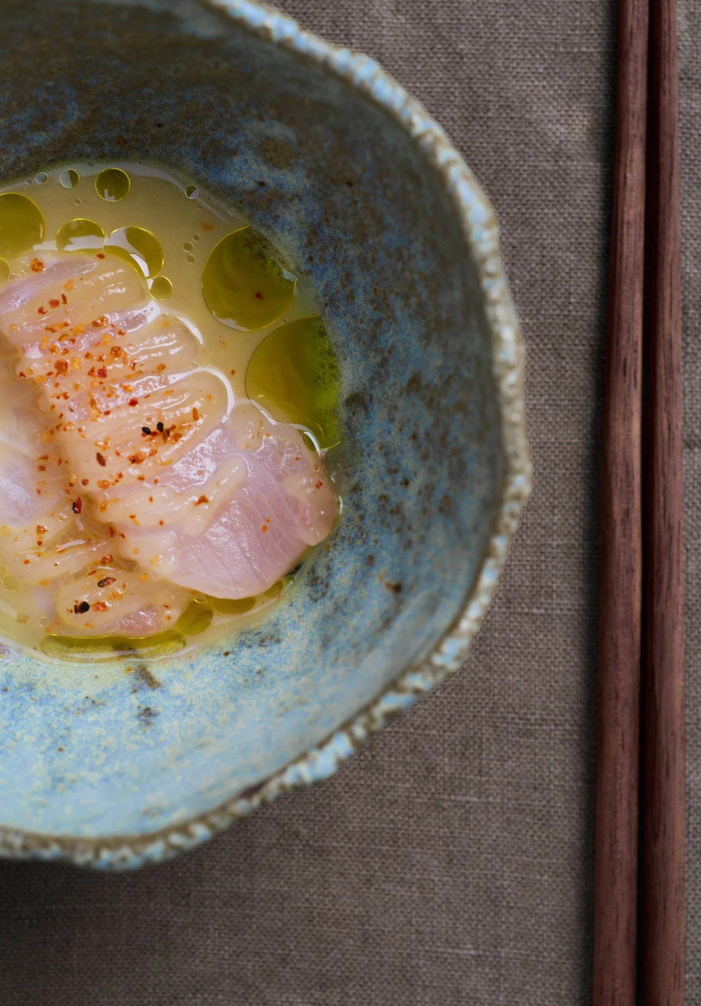 A ceramic bowl with a textured rim contains raw fish slices topped with chili flakes, surrounded by green oil or sauce, placed on a woven fabric with chopsticks beside it.