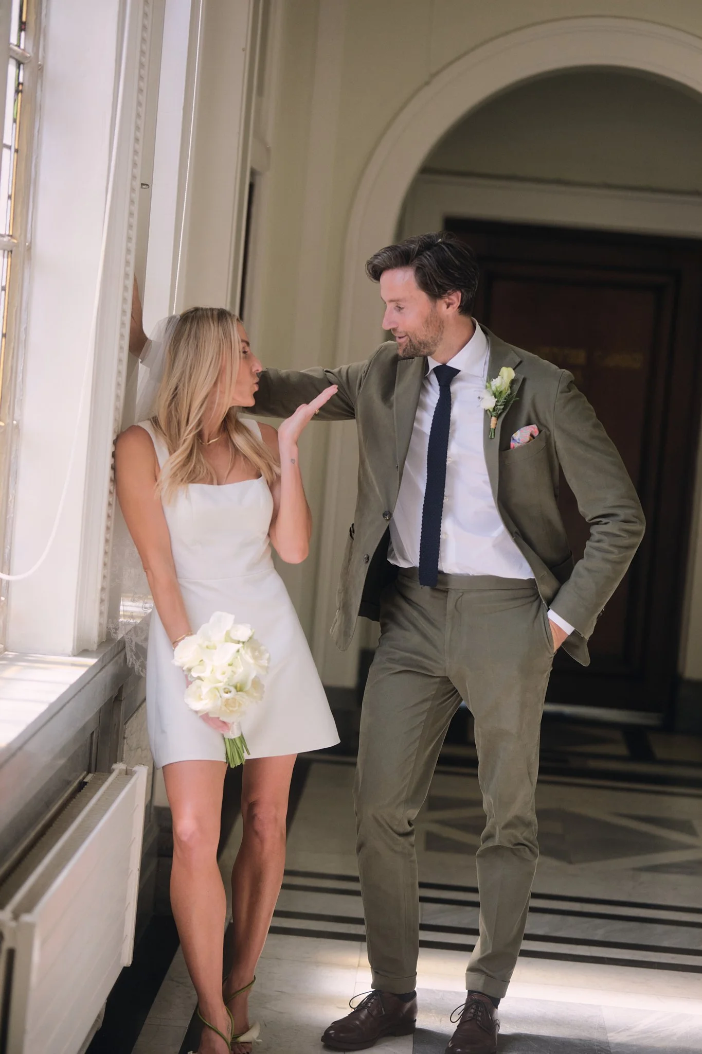 A bride and groom share a moment in a hallway. The bride, in a white dress and heels, holds a bouquet of white flowers. The groom, in a gray suit, white shirt, and dark tie, has a boutonniere on his lapel. They are close, smiling, and gazing at each 