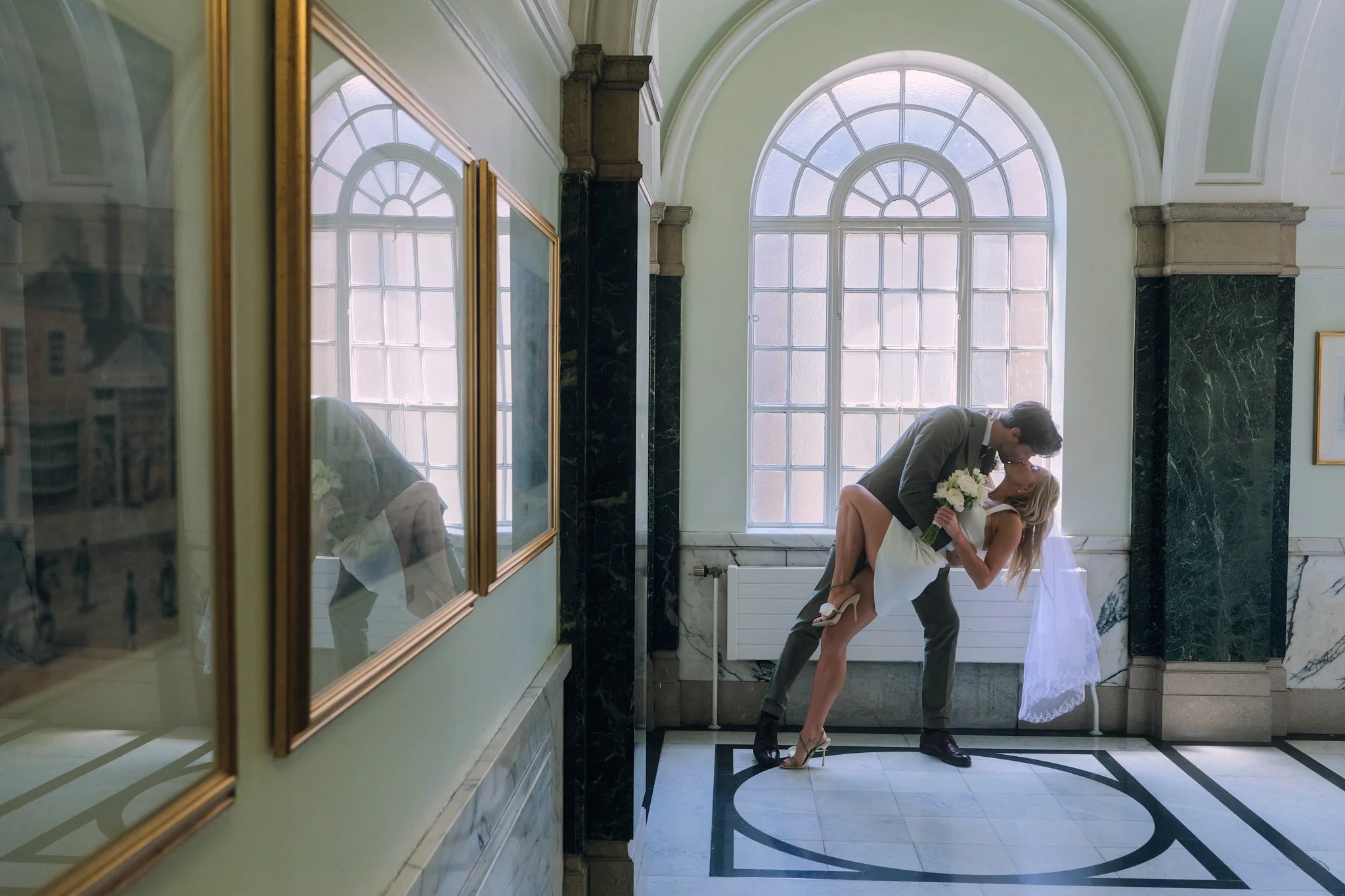 A man and woman in wedding attire sharing a kiss in front of large arched windows in a bright, elegant room with black and white marble floors and decorative columns.