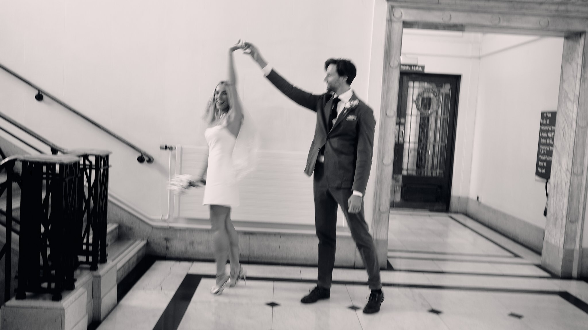 A couple dancing on a staircase in a building, with the woman wearing a white dress and heels, and the man in a suit.