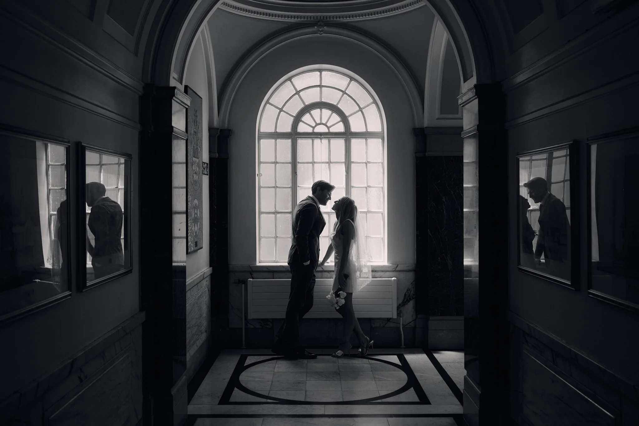 A black and white photo of a man and woman facing each other in front of a large window with an arched top. They are holding hands and looking into each other's eyes, with the woman holding a bouquet of flowers. The scene appears to be inside a build
