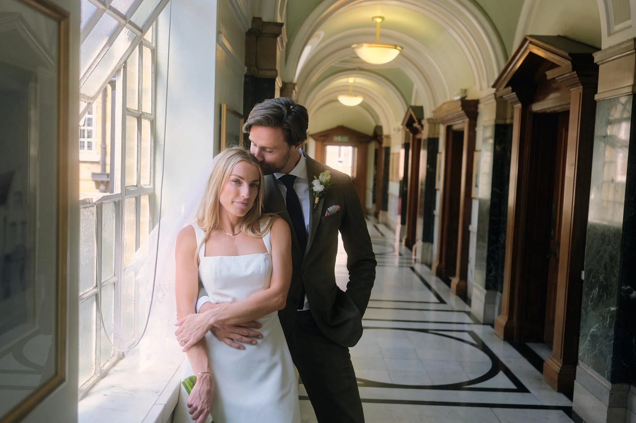 A couple dressed in wedding attire in a long, elegant corridor with arched ceilings and large windows, sharing a tender moment.