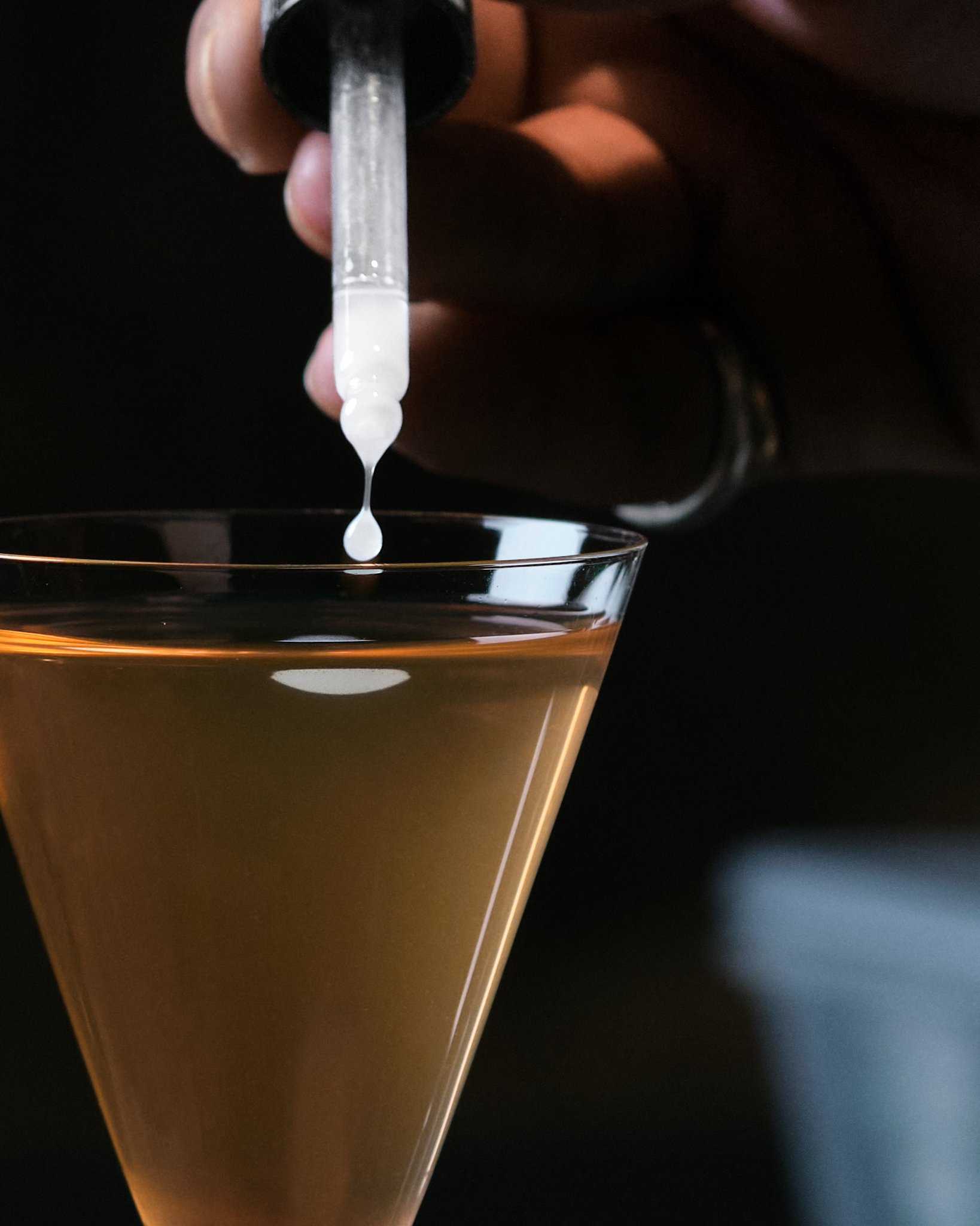 A clear dropper pipette releasing a drop of a clear liquid into a glass container with a dark background.