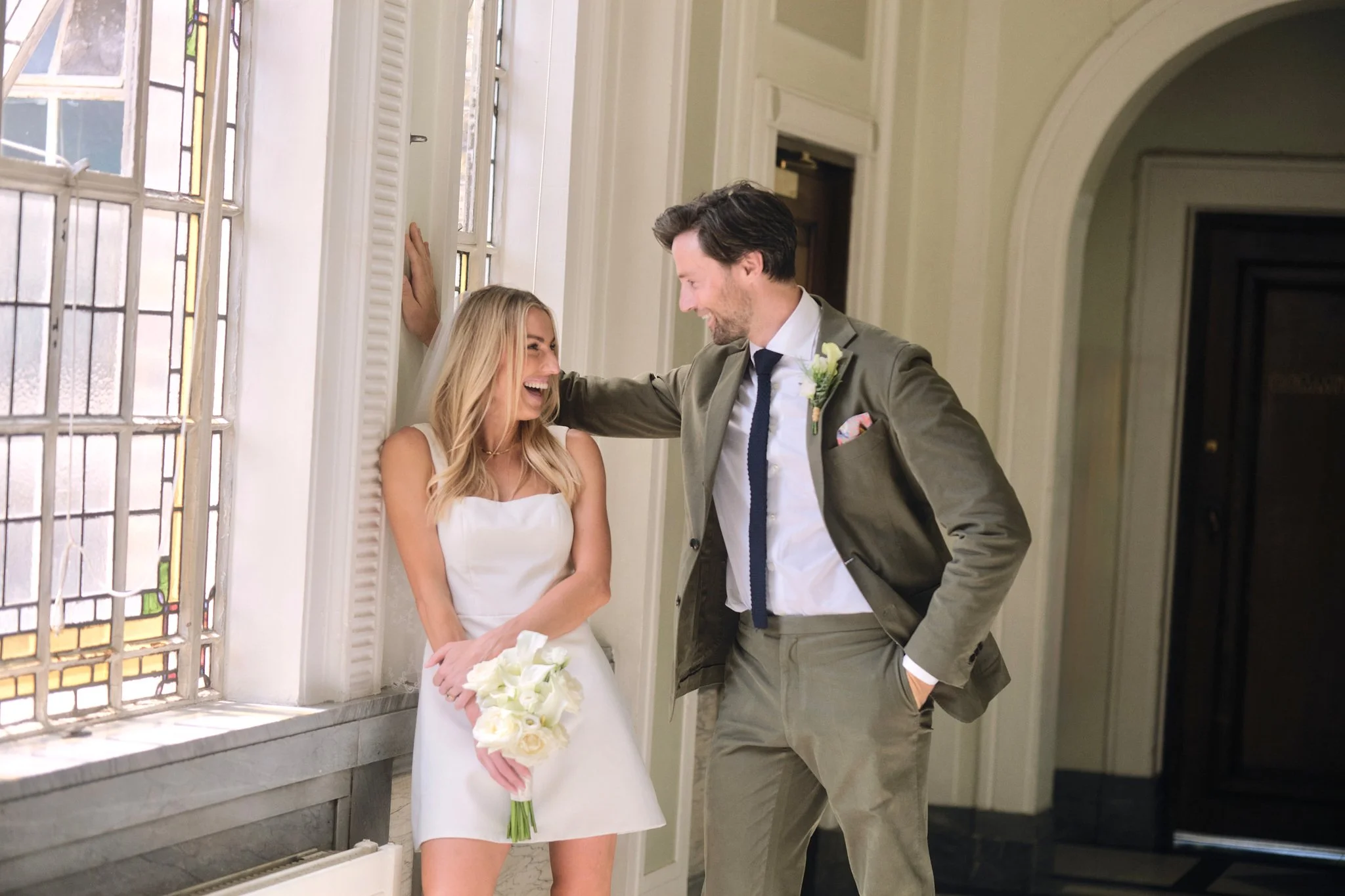 A bride and groom sharing a joyful moment inside a building next to a stained glass window. The bride is holding a bouquet of white calla lilies and is leaning against the window, while the groom, dressed in a suit with a boutonniere, is smiling and 