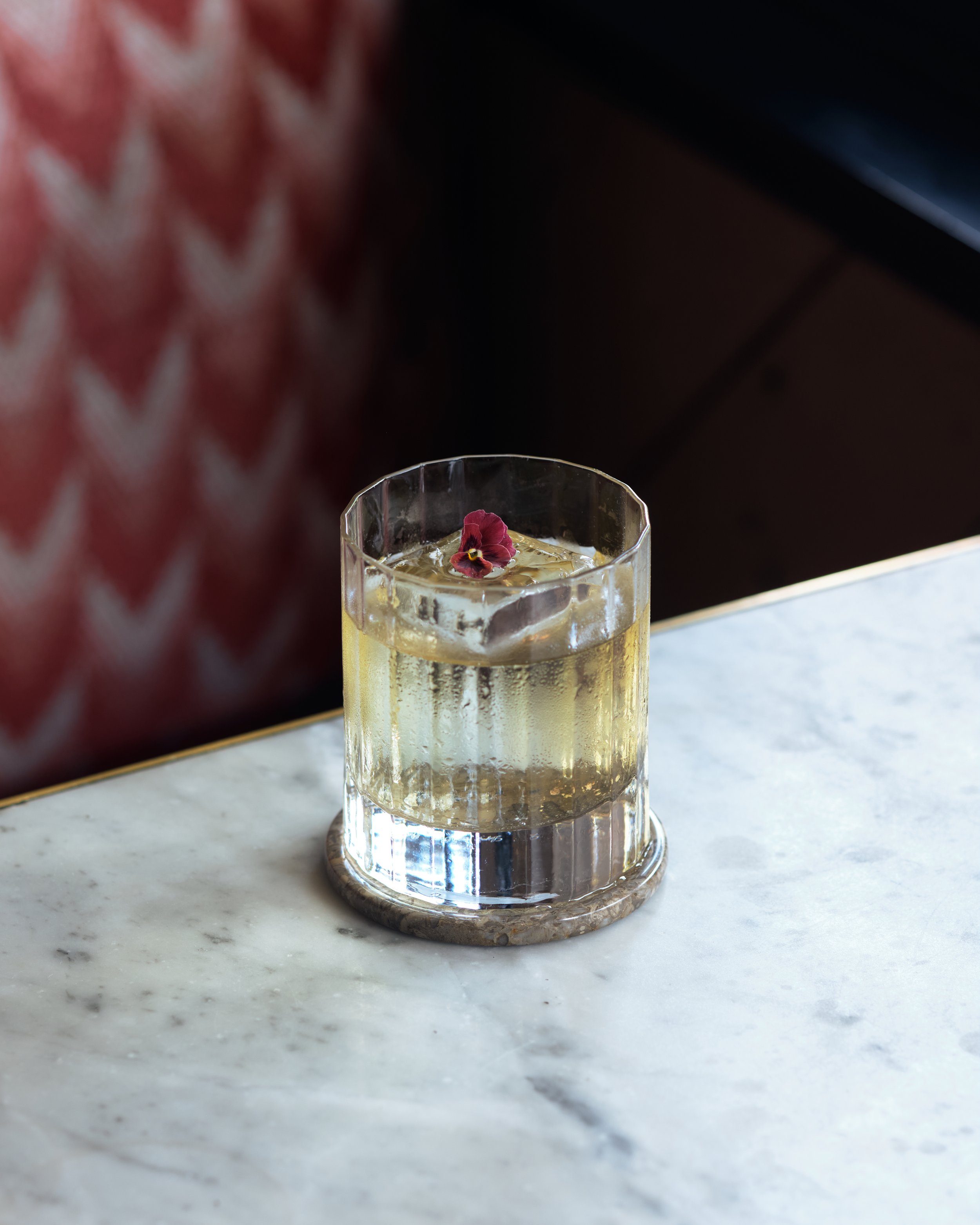 A clear glass filled with a light-colored beverage and ice, garnished with a small edible flower, placed on a marble surface.