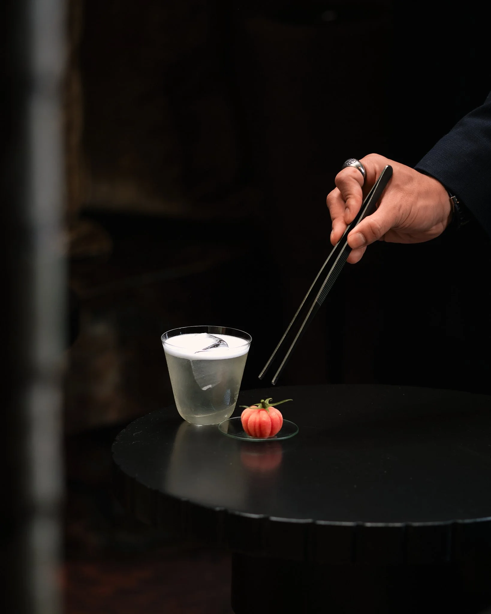 Hand holding chopsticks over a martini glass with ice cubes, a small pink checkered tomato, and a sprig of rosemary on a black surface with a dark background.