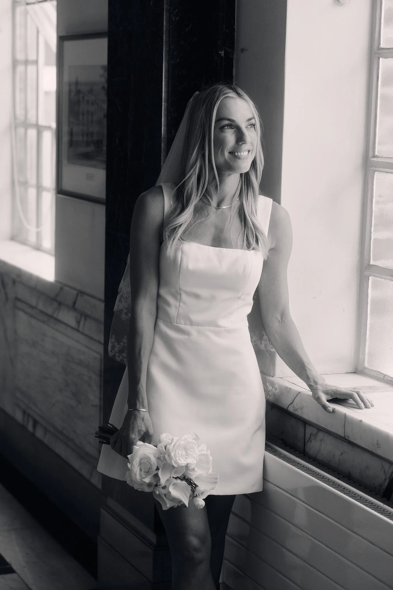 Black and white photo of a woman in a wedding dress, holding a bouquet of white roses, standing by a window with her hand resting on the windowsill, smiling and gazing out.
