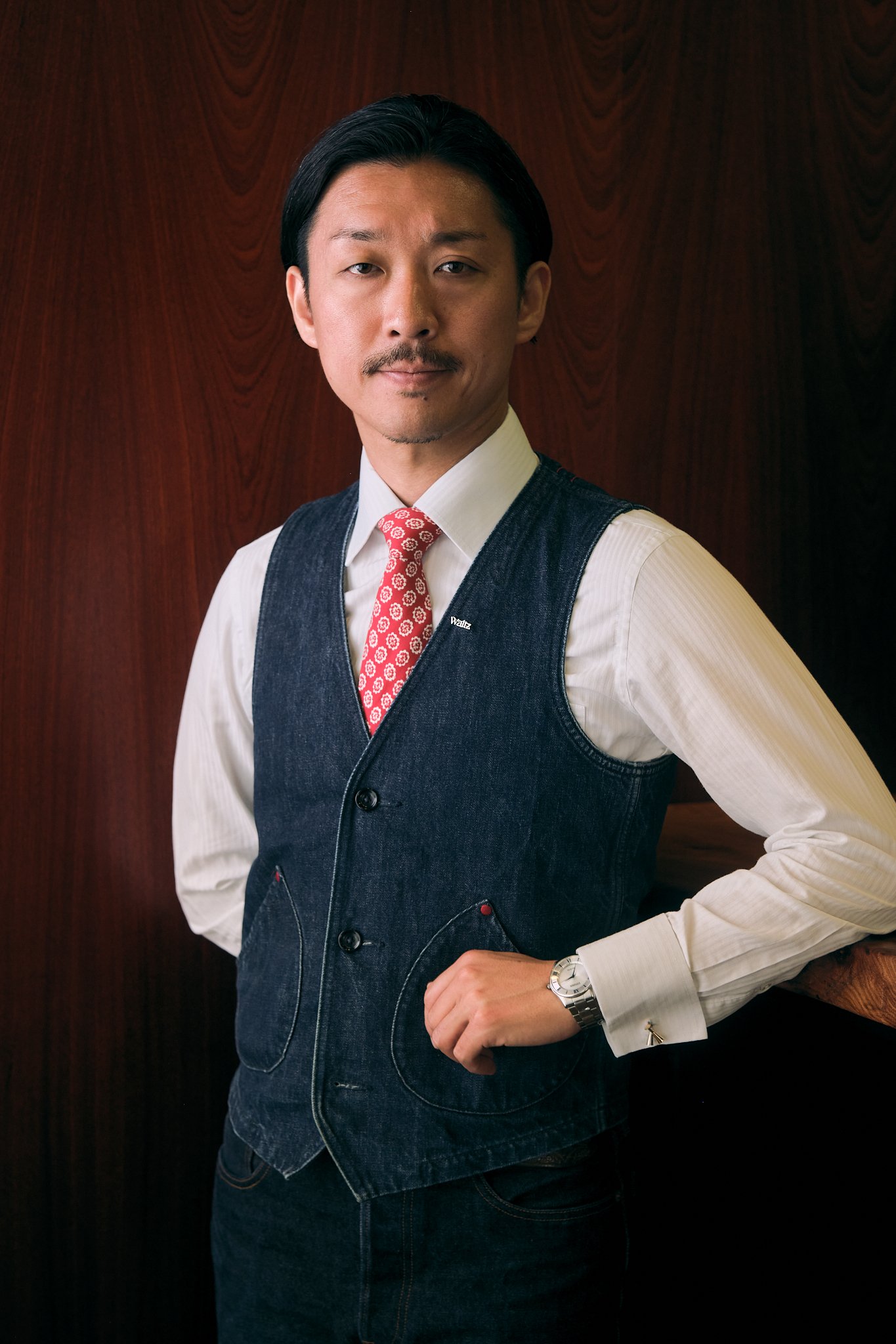 A man with black hair, wearing a white shirt, red patterned tie, denim vest, dark blue jeans, a wristwatch, and cufflinks, standing against a wood-paneled wall.
