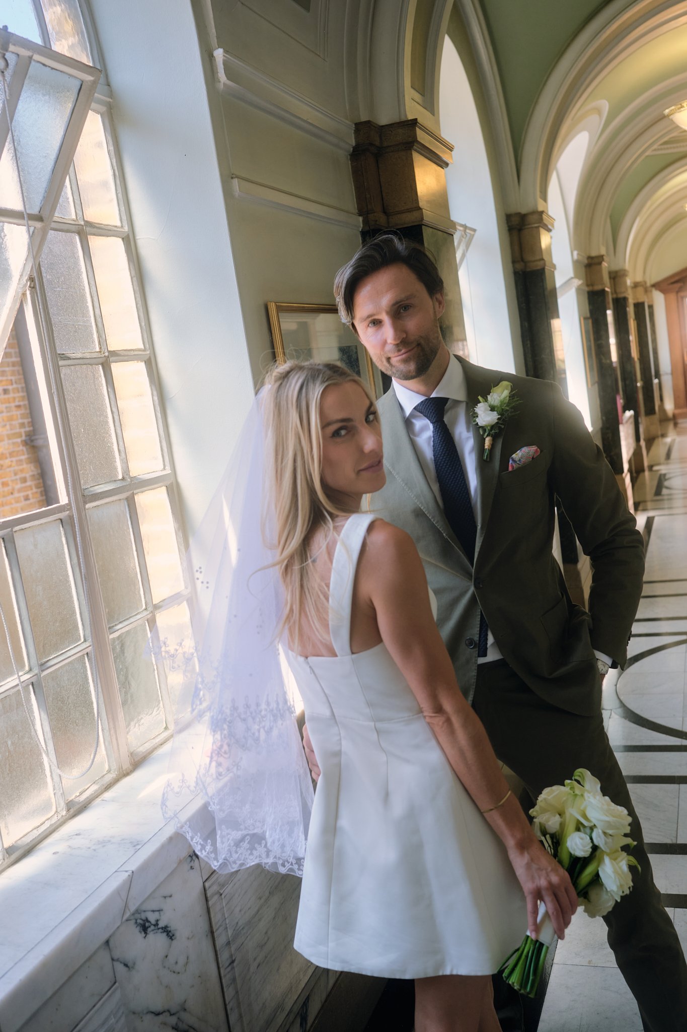 A bride and groom standing inside near large windows and marble wall, smiling for a wedding photo, with the bride holding a bouquet of white flowers.