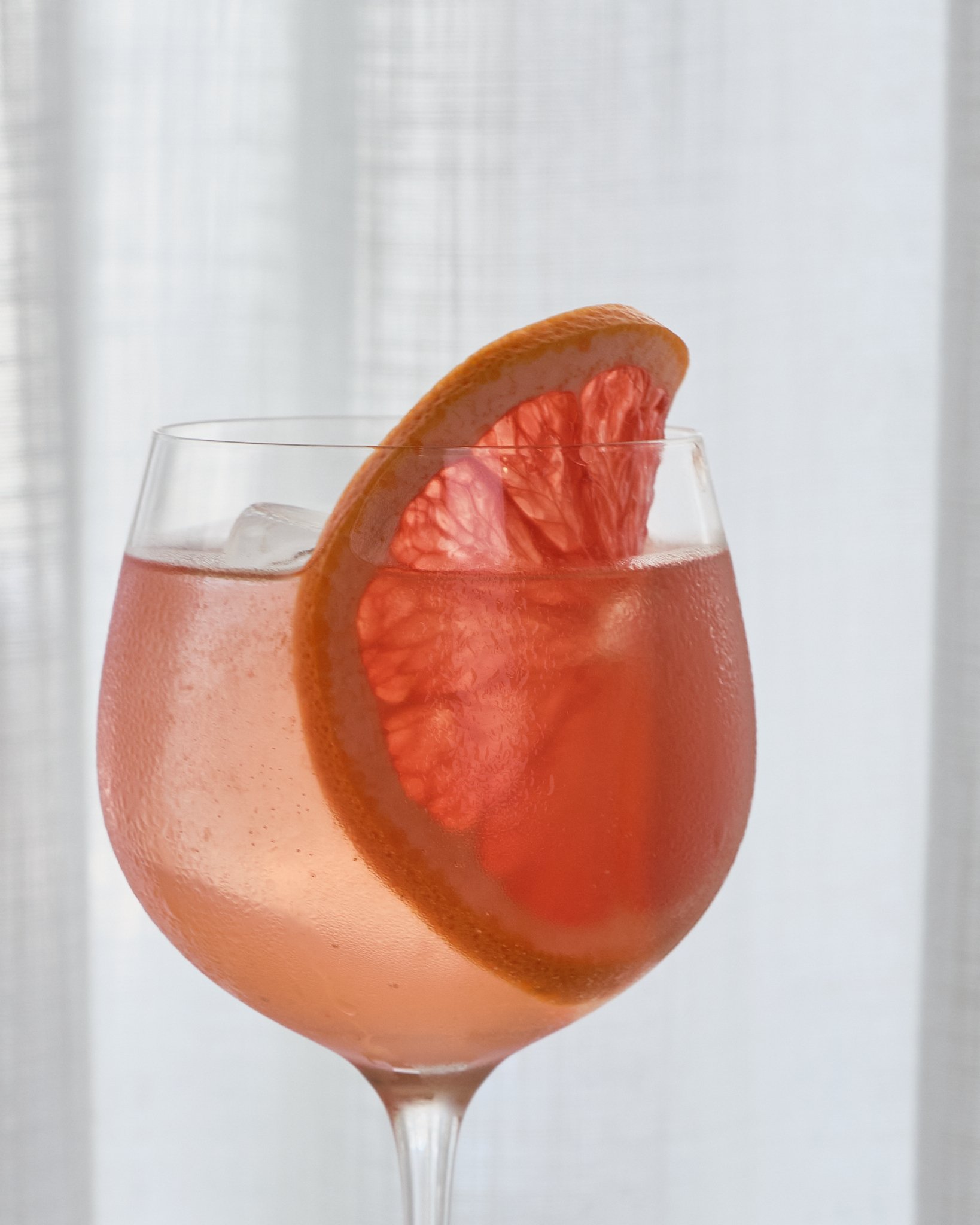 A pink grapefruit cocktail garnished with a slice of grapefruit in a stemmed glass, with ice cubes inside.