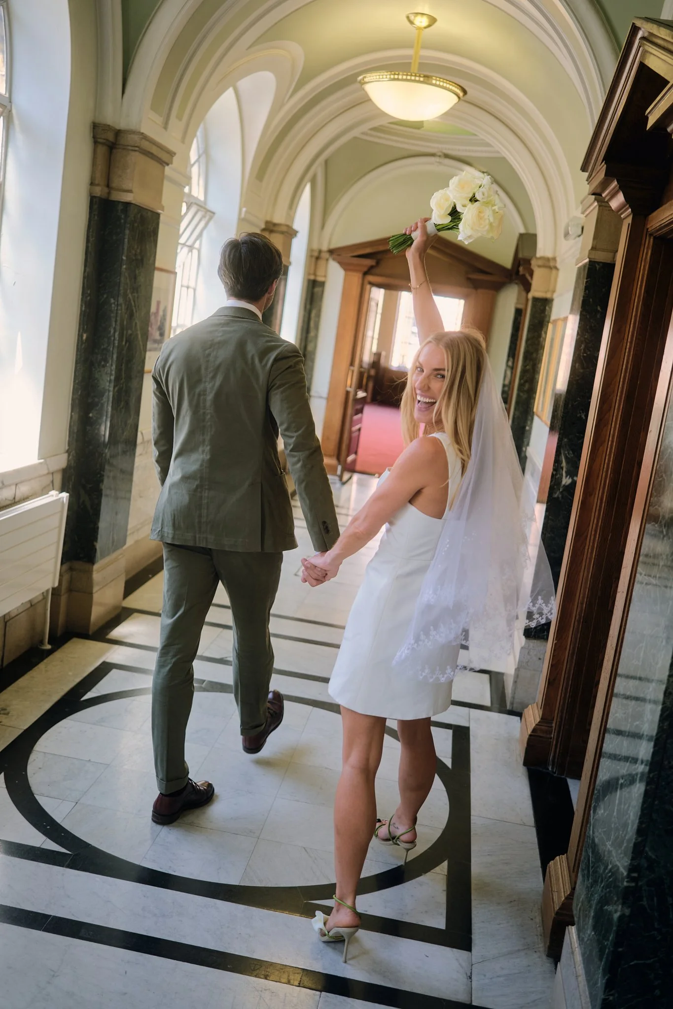 A smiling blonde woman in a white sleeveless dress and high heels holding a bouquet of white roses raises her arm in celebration as she walks hand-in-hand with a man in a suit through a historic hall with arched ceilings and marble columns.