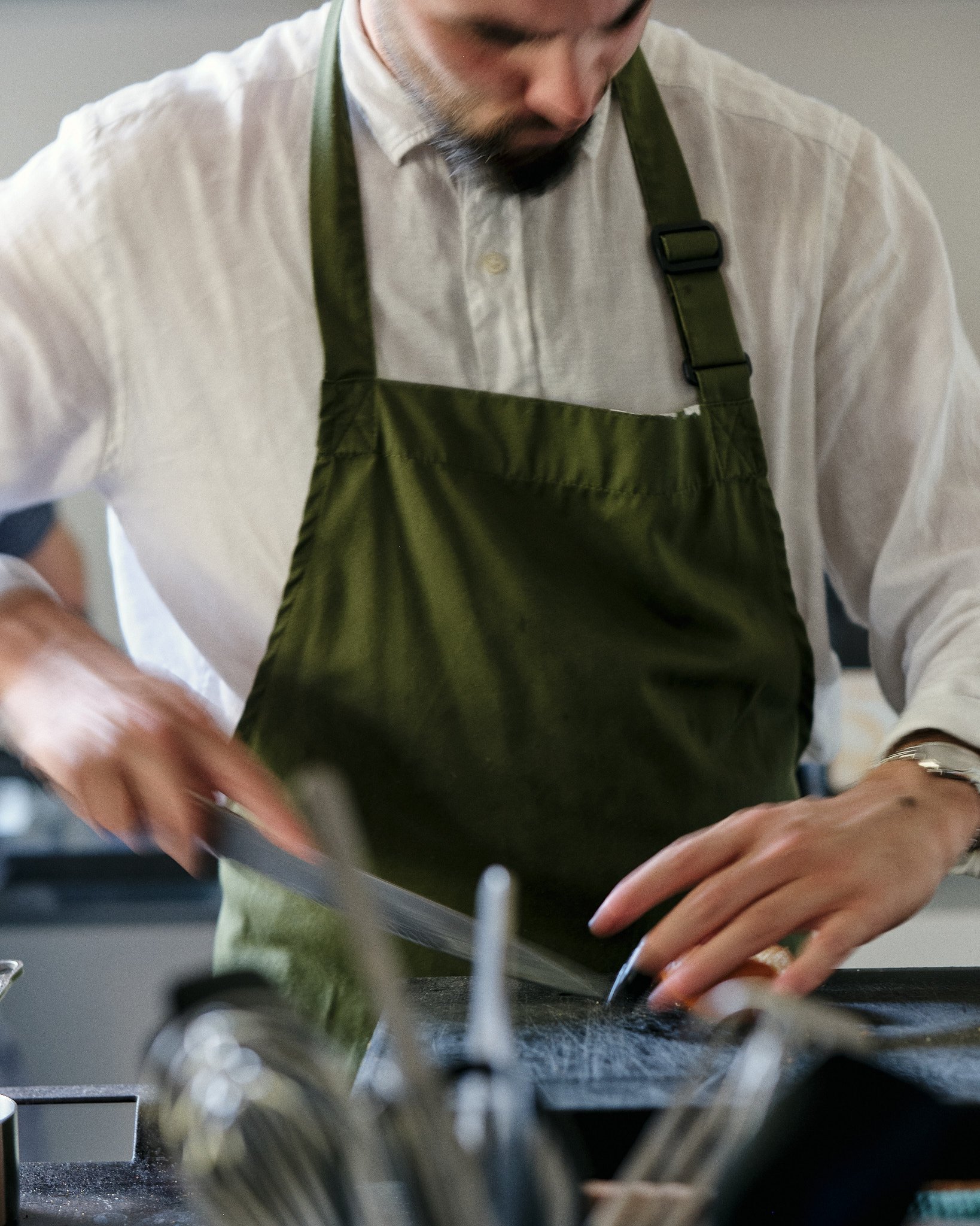 A man in a white shirt and green apron is working at a kitchen counter, focusing on task with his hands near the edge of the counter.
