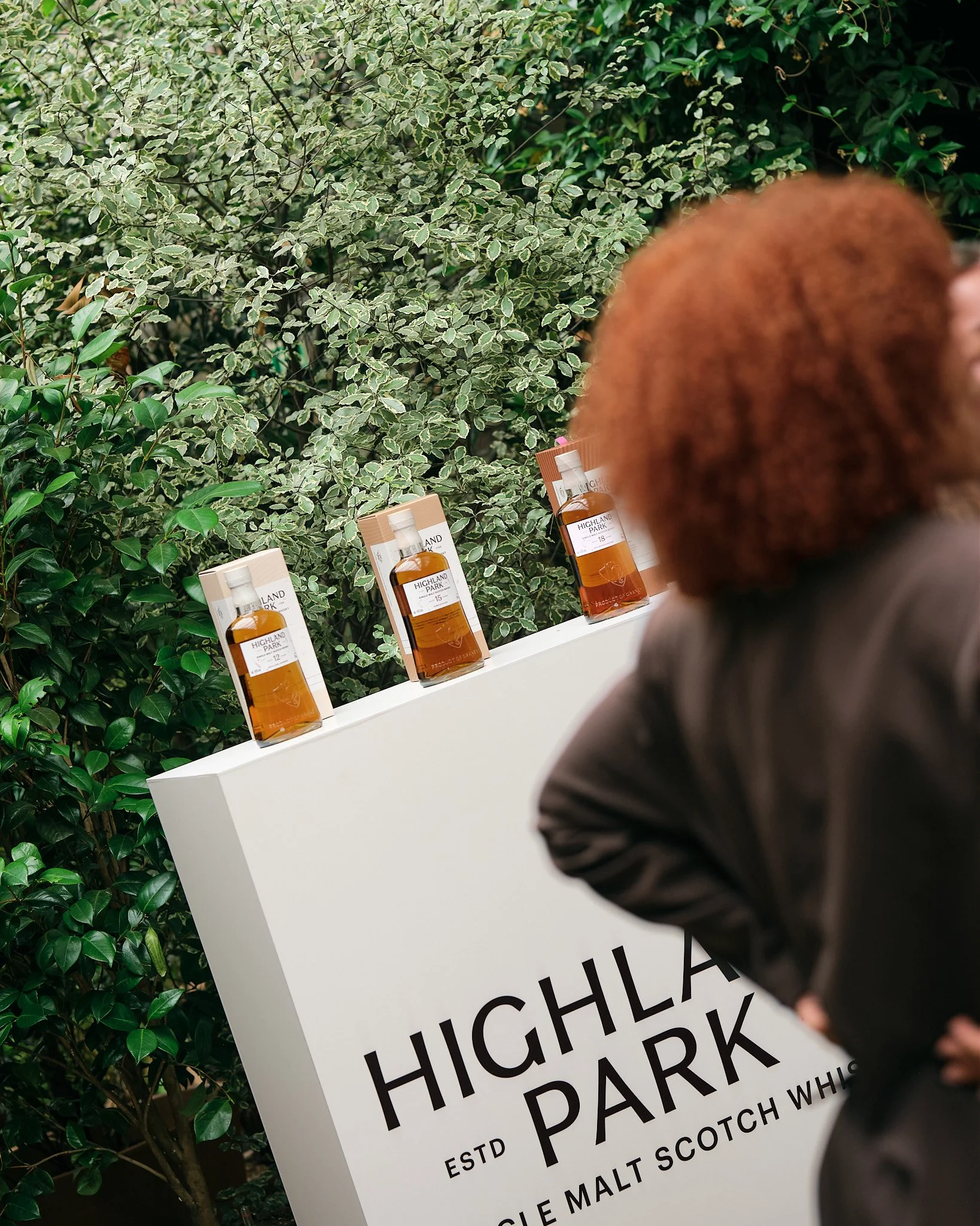 Close-up of a woman with red hair next to a white sign with black and gray text, and bottles of Highland Park whisky, set against a background of green leafy plants.