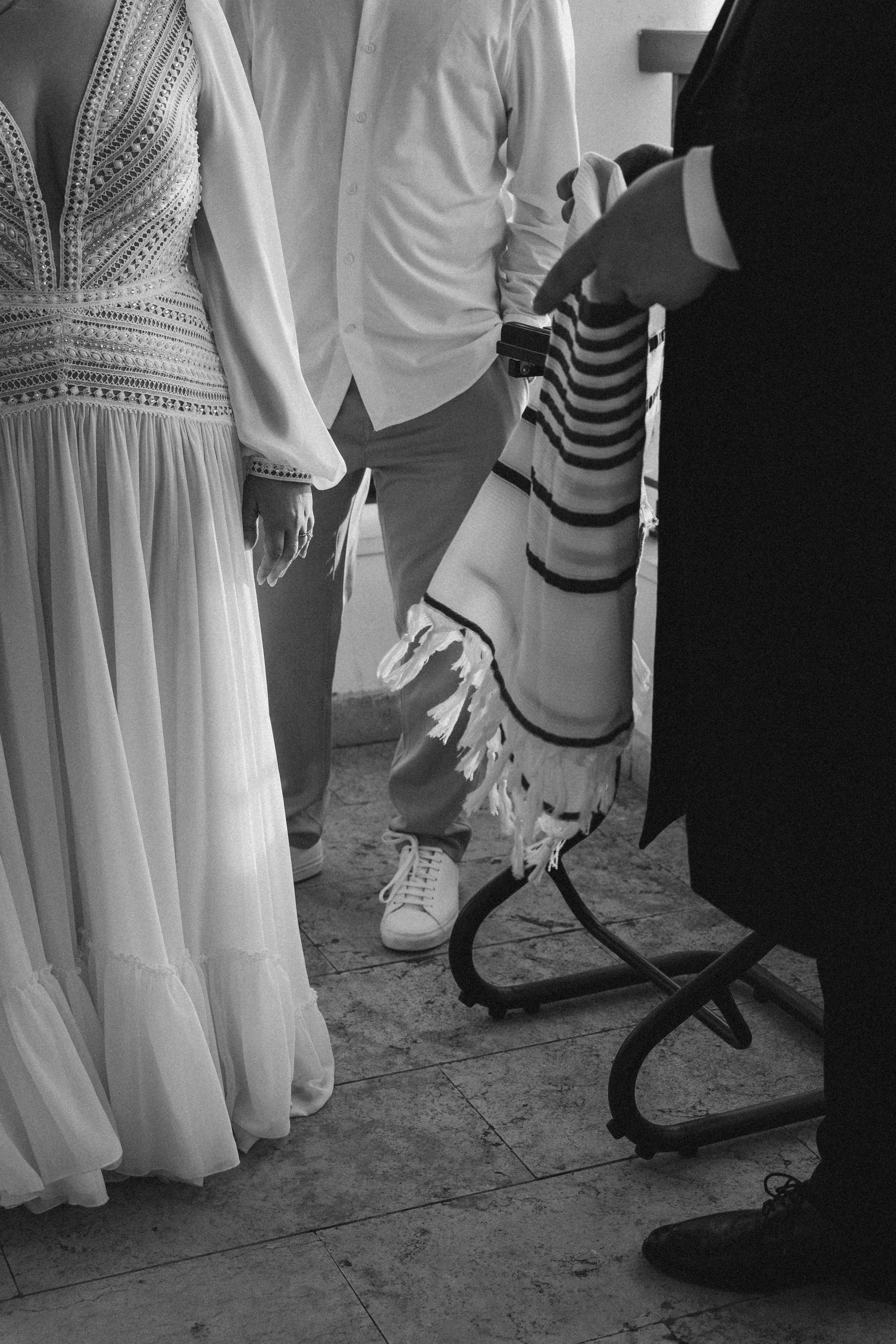 Black and white photo of people at a wedding or formal event, showing part of a bride's white dress with intricate lace details, a person in sneakers and trousers, and a man in a suit with a striped cloth on his arm, seated on a chair.